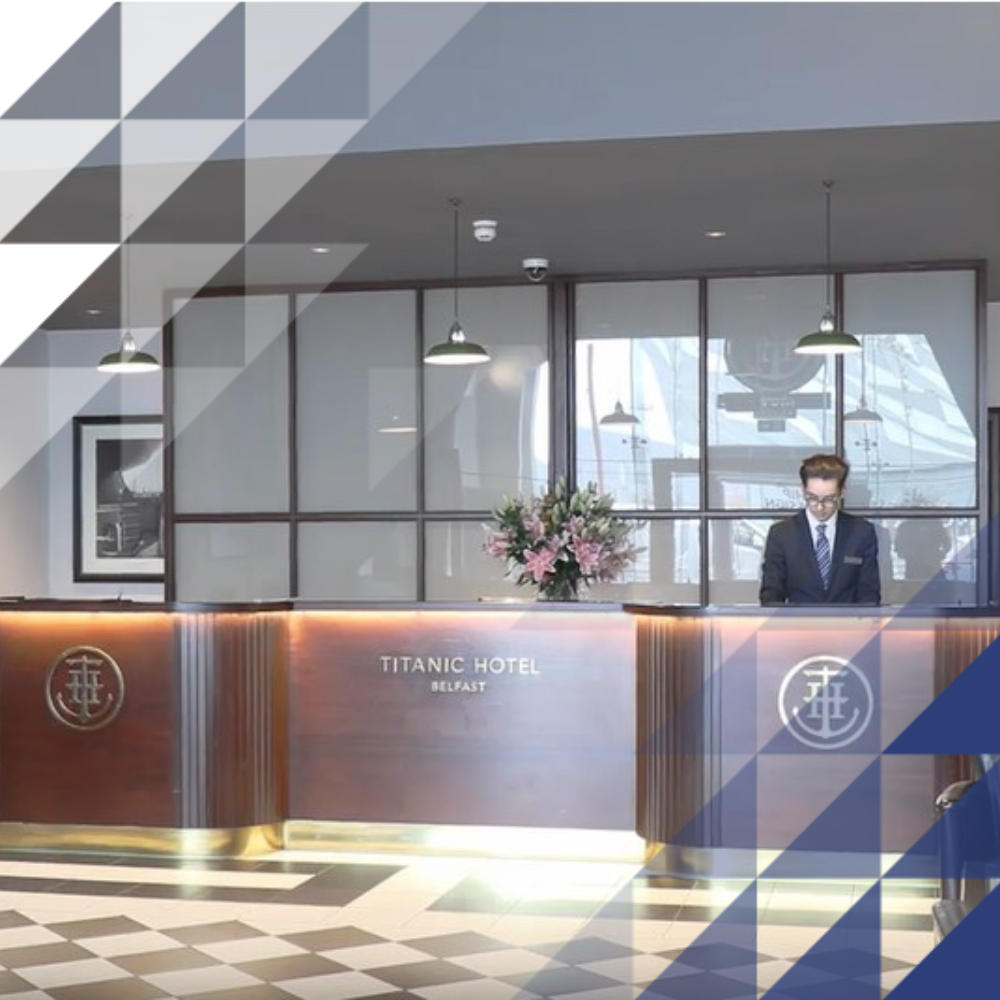 Titanic Hotel Belfast reception with a polished wood and brass counter, monochrome tiled floor, maritime artwork, and a staff member on duty.
