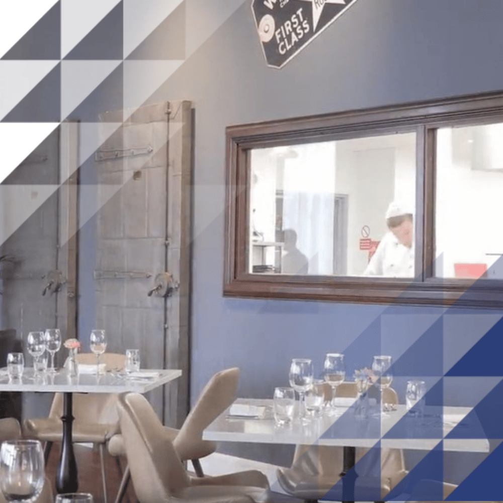 Restaurant dining area with white table settings, wine glasses, and tan chairs, featuring a large window into the kitchen where a chef in a white uniform is working, a 
