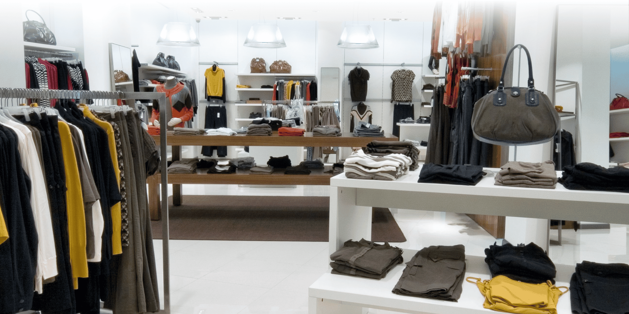 Boutique di abbigliamento contemporaneo con capi appesi alle grucce, vestiti piegati e borse esposti sui tavoli, e scaffali illuminati sullo sfondo.