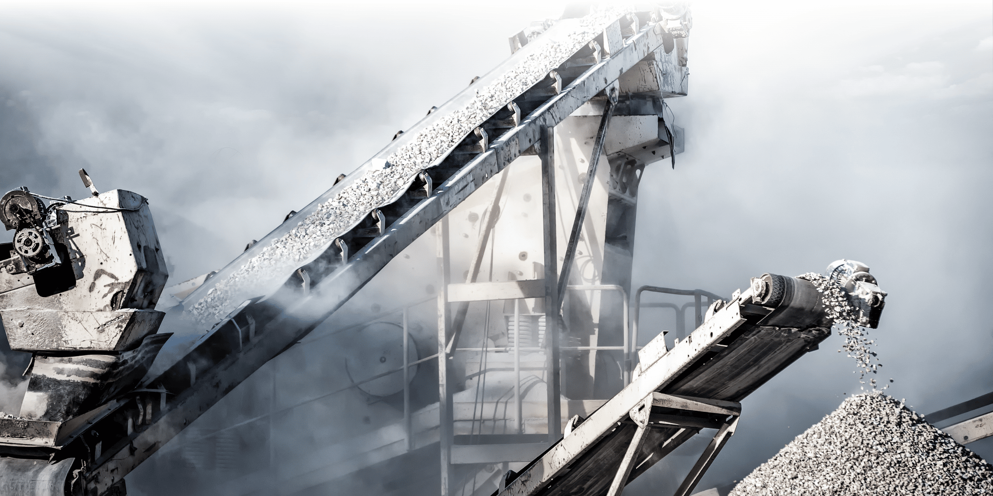 Industrial conveyor belts carrying crushed aggregate at a quarry or processing plant, with dust rising against a dramatic cloudy sky.