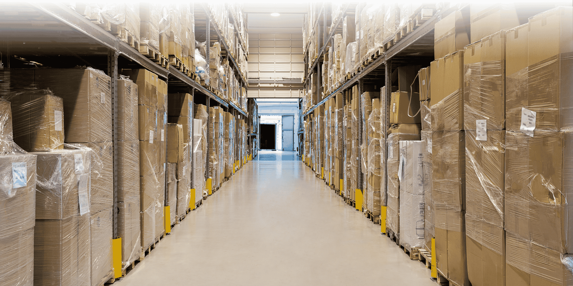 A narrow warehouse aisle flanked by shelving stacked with cardboard boxes and wrapped pallets, leading to a loading bay door at the far end.