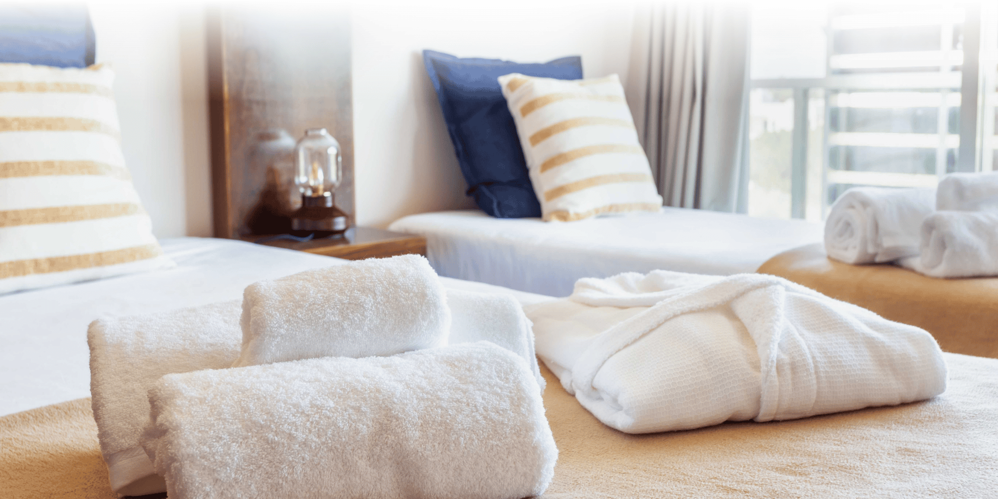 wo beds in a hotel room with towels and sheets layed on top. Both beds contain a beige cover, stripey and dark blue pillows