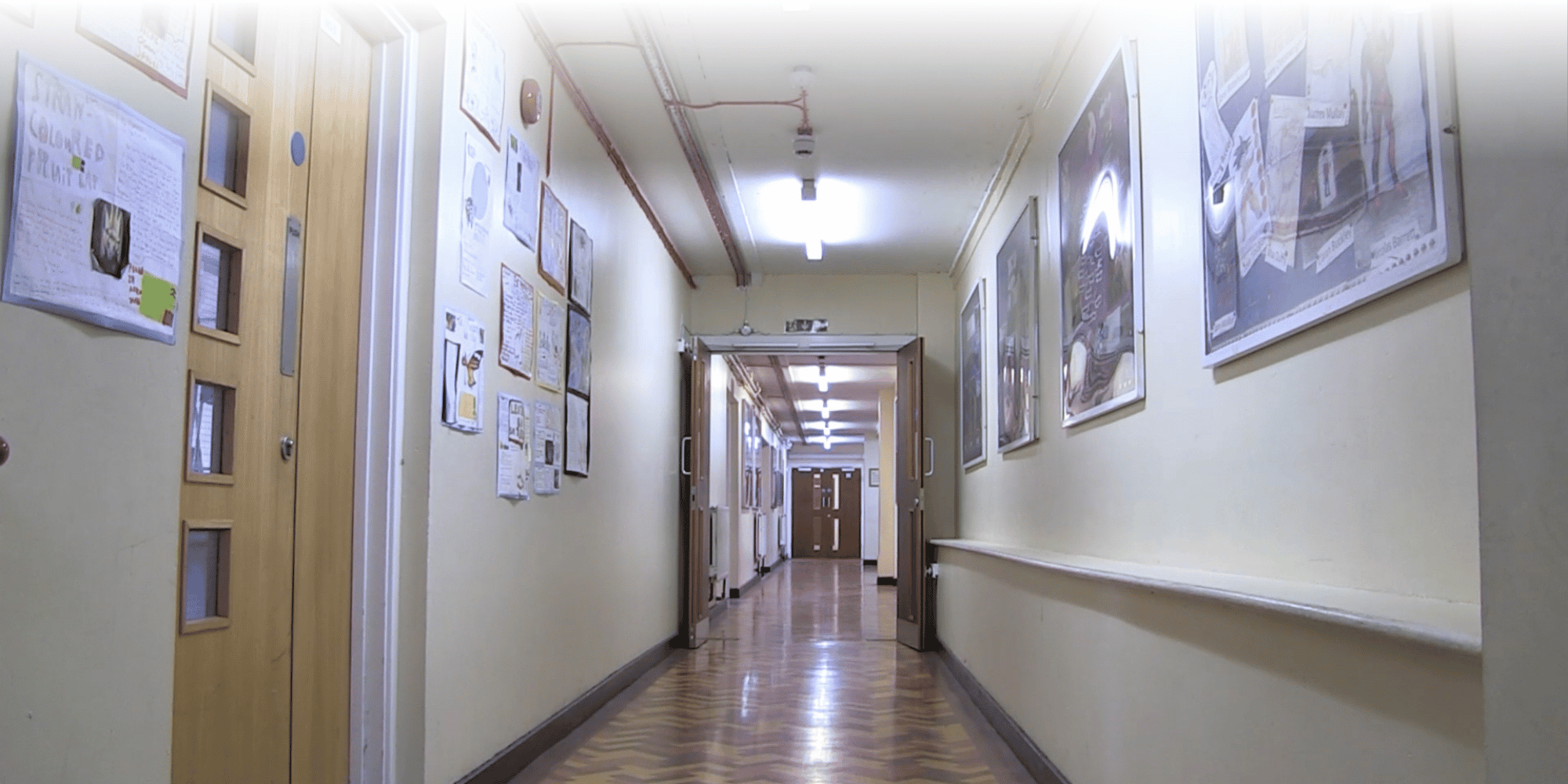 School corridor with cream walls displaying children's artwork, a red fire alarm pipe along the ceiling, and fluorescent strip lighting.