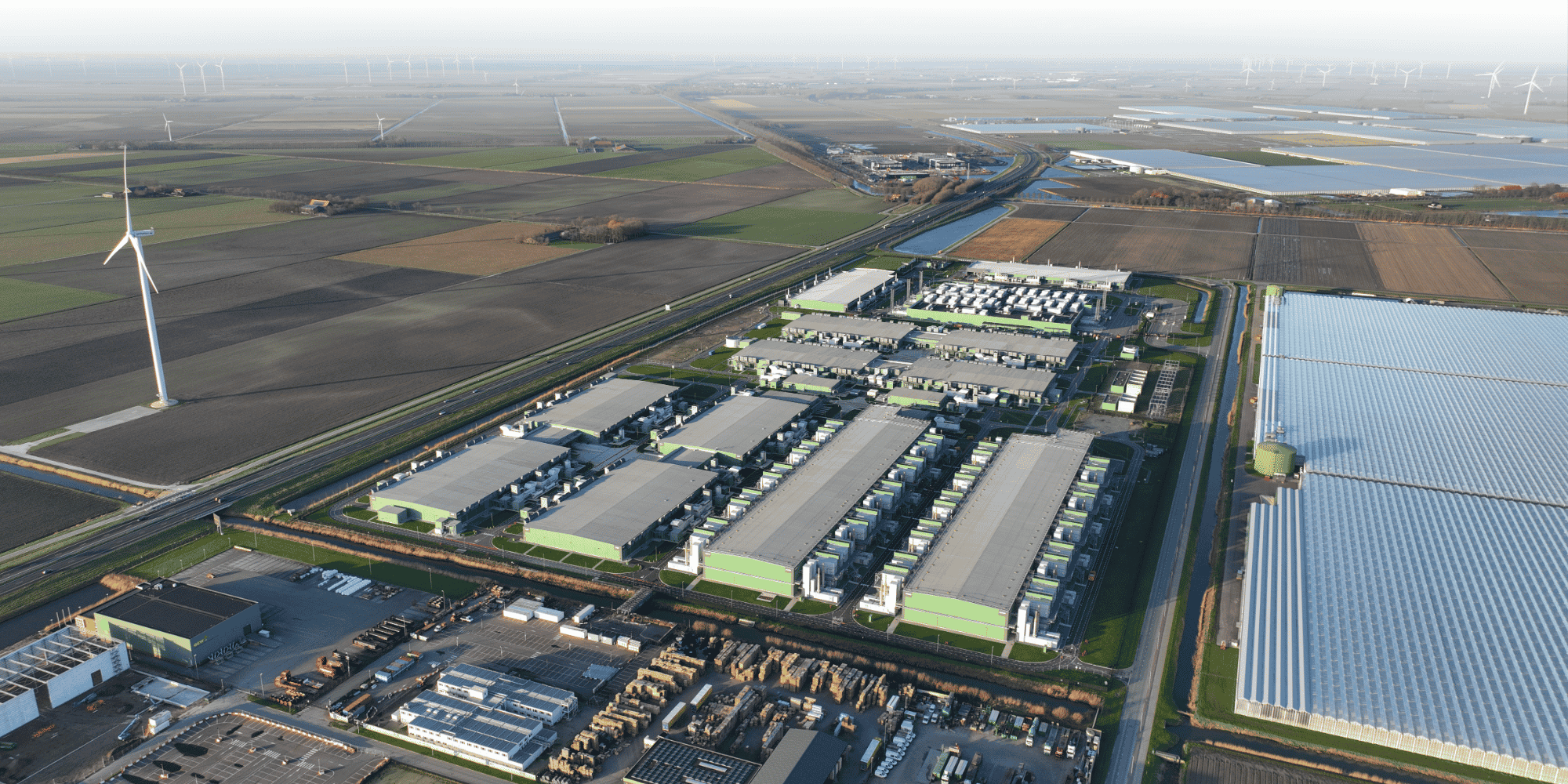 Bird's-eye view of industrial estate with rows of warehouse units, bordered by open fields, wind turbines and a solar farm under a clear sky.