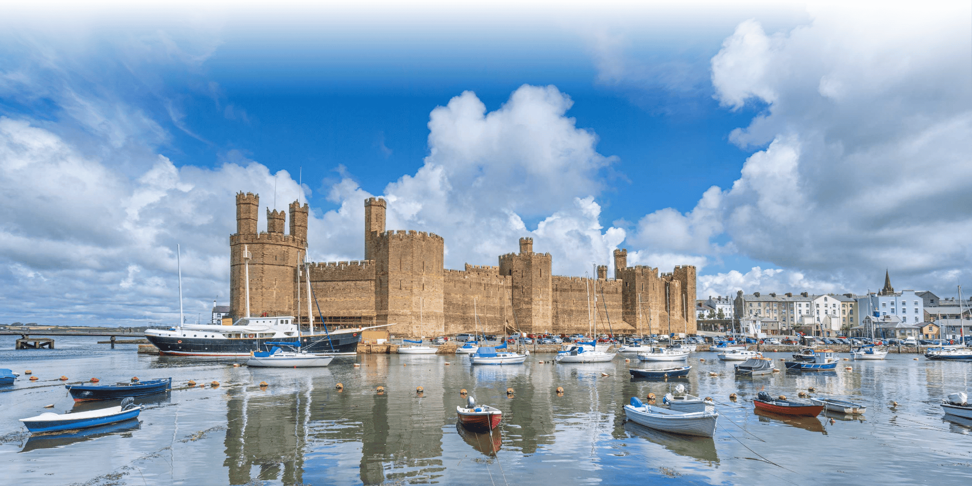 Caernarfon Castle's medieval sandstone walls and towers reflected in the harbour, with moored sailing boats under a partly cloudy sky.