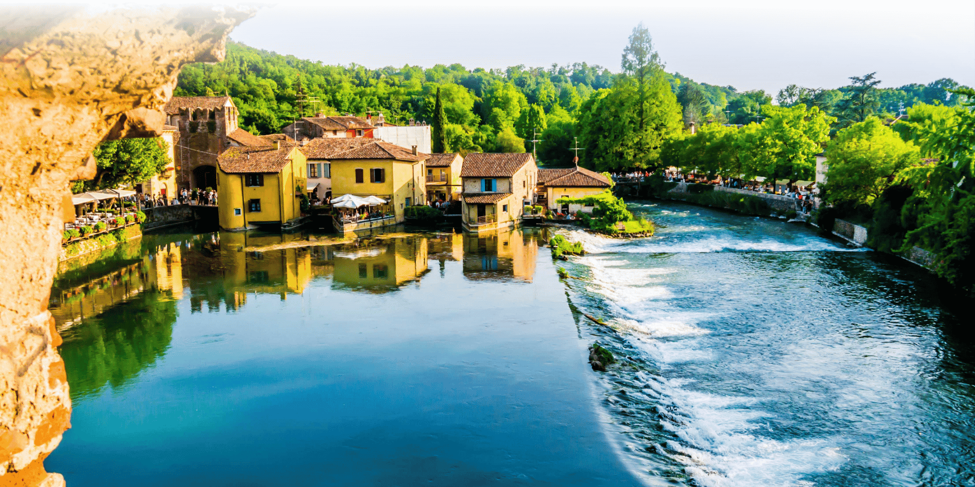 Edifici in pietra e un mulino giallo che costeggiano un fiume tranquillo con una diga, sullo sfondo delle verdi colline di Borghetto sul Mincio, in Italia.