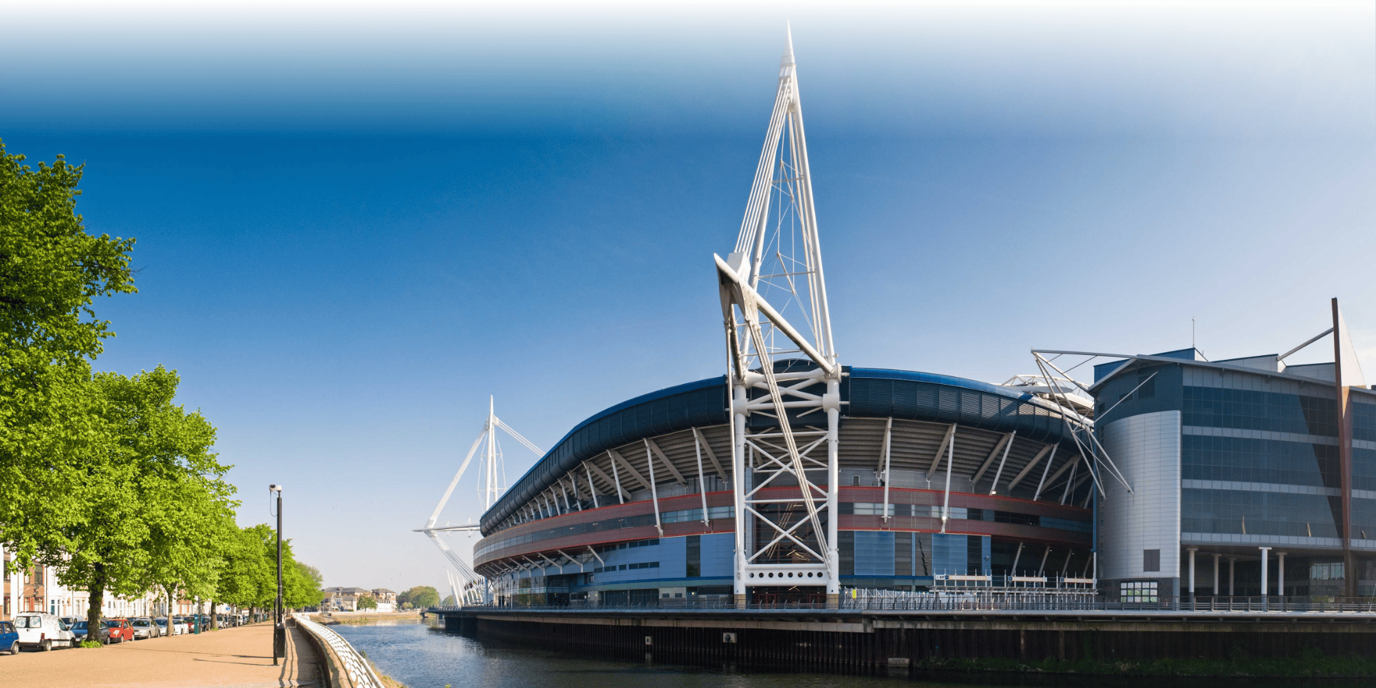 Exterior view of the Principality Stadium in Cardiff, seen from the riverside on a clear sunny day, with its distinctive white steel masts and retractable roof structure visible against a blue sky.