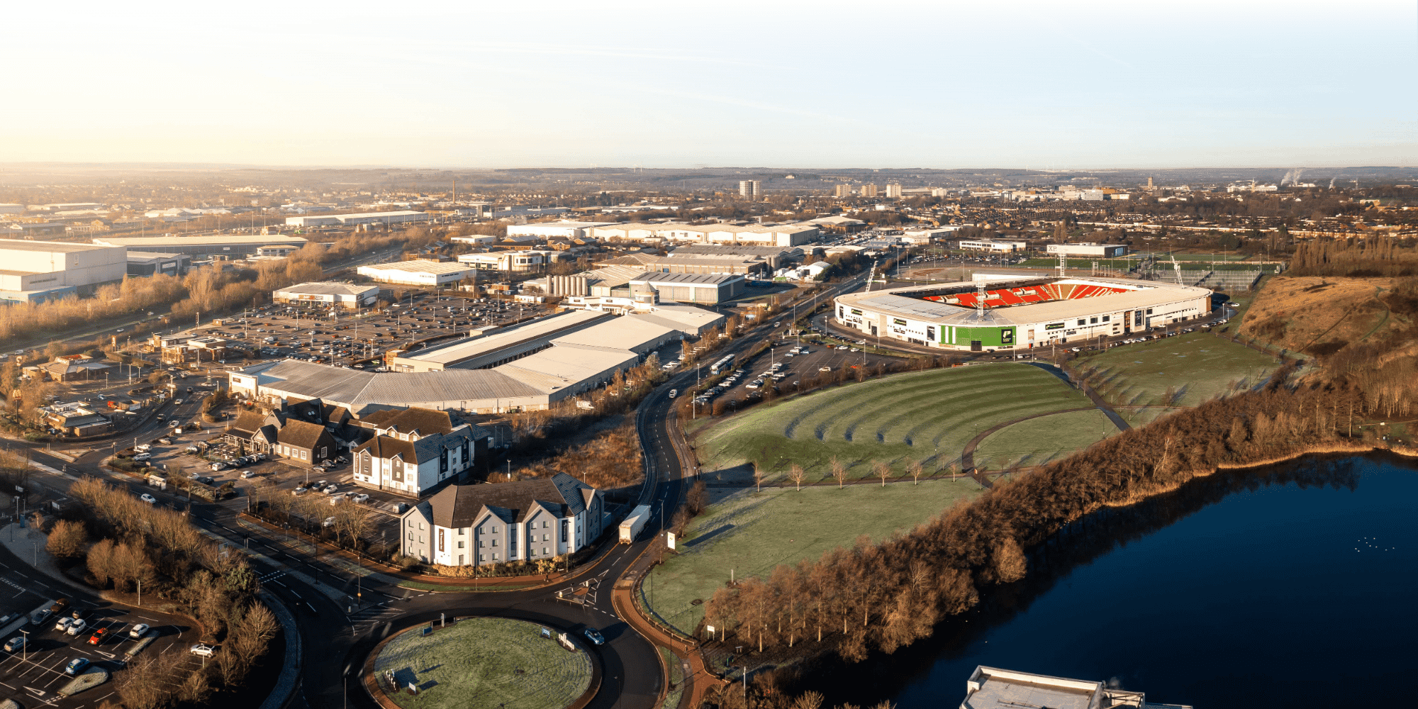Aerial winter view of an urban area showing a red-and-green football stadium, retail park, hotel, roundabout, and lake, with the town skyline beyond.