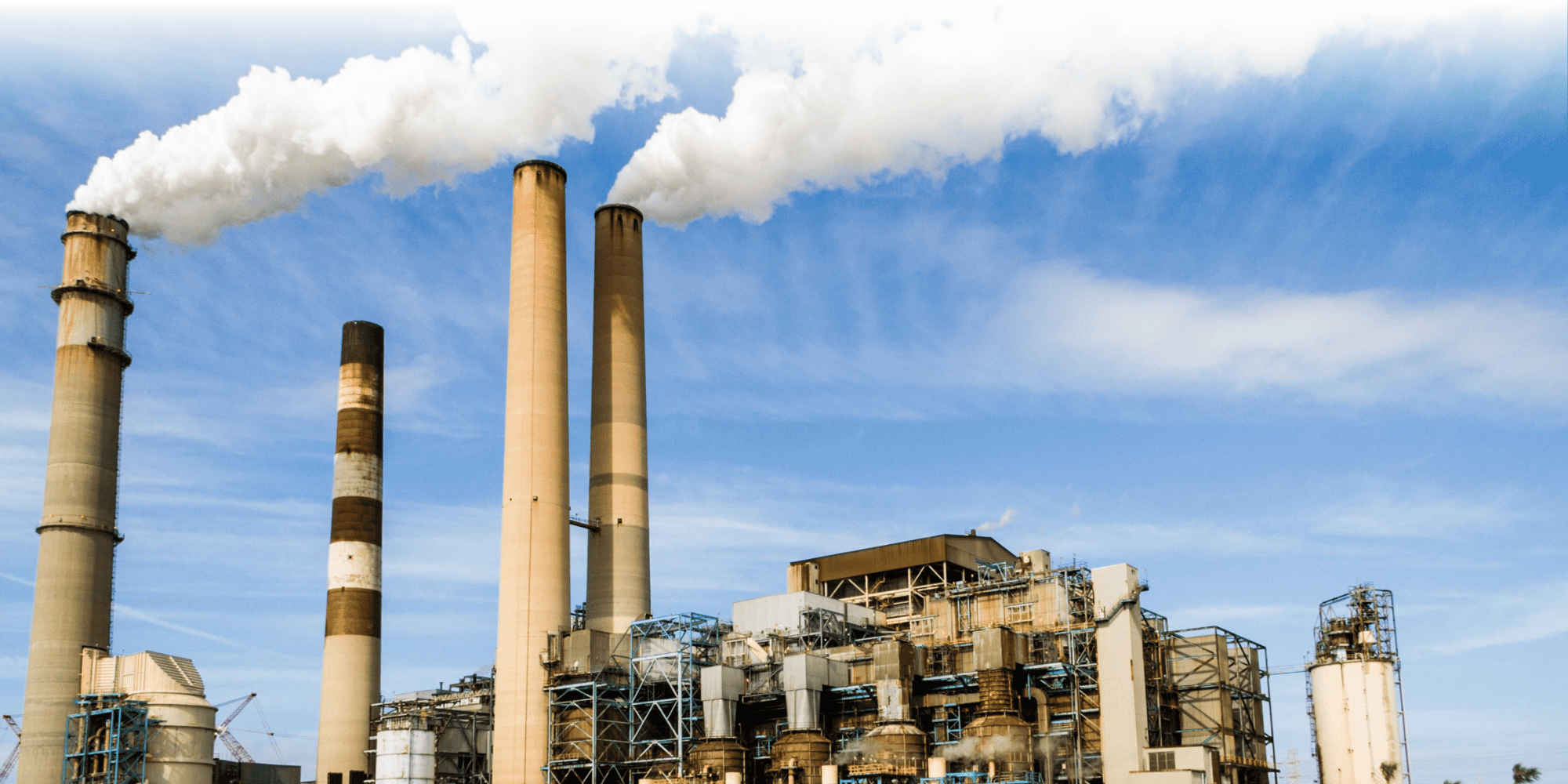 Industrial facility with four smokestacks emitting white smoke, surrounded by pipework, storage tanks, and processing equipment.