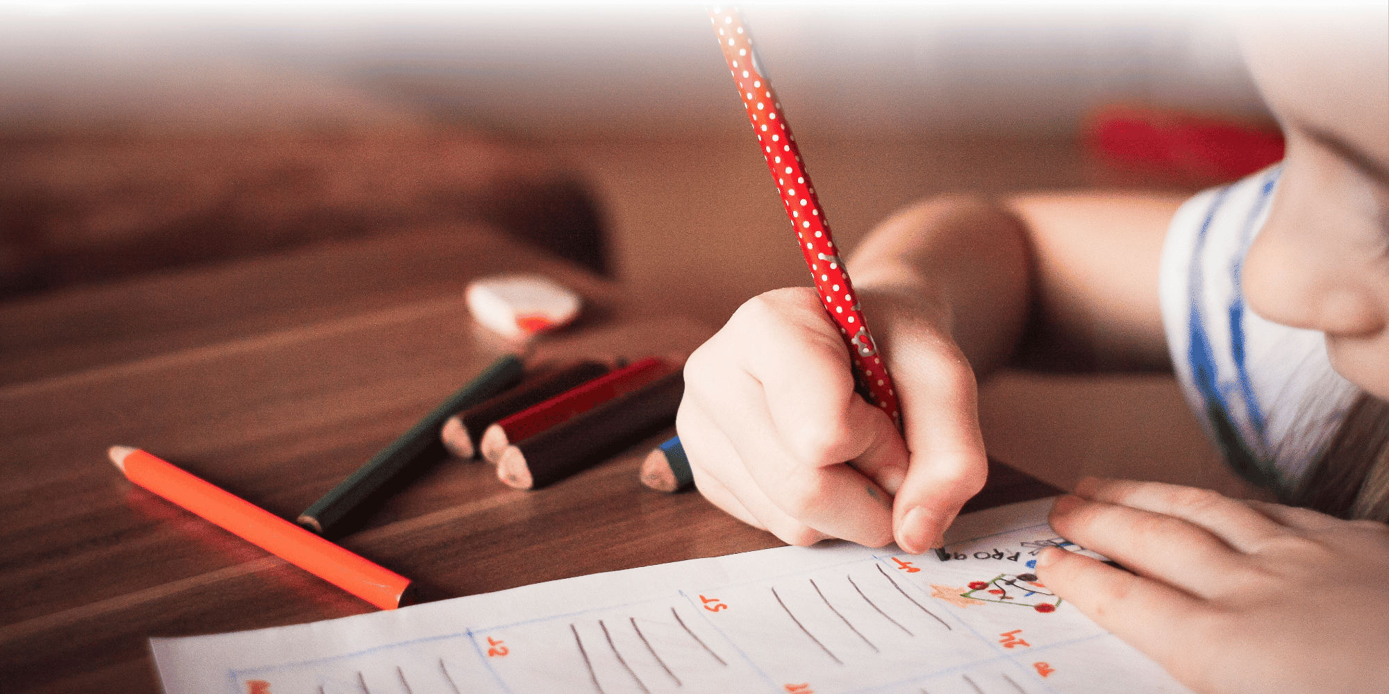 Close-up of a child's hand writing with a red polka-dot pencil on a lined worksheet with numbered rows and drawings, with colored pencils on the table nearby.