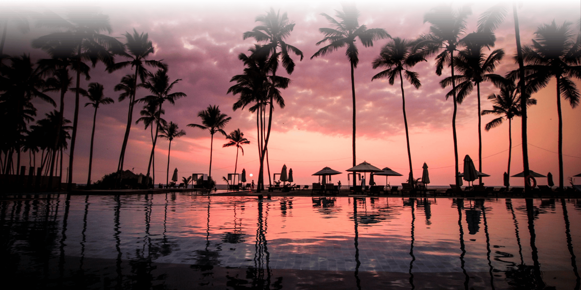 A luxury hotel infinity pool at sunset, with silhouetted palm trees and parasols reflected in the still water against a dramatic pink and purple sky.