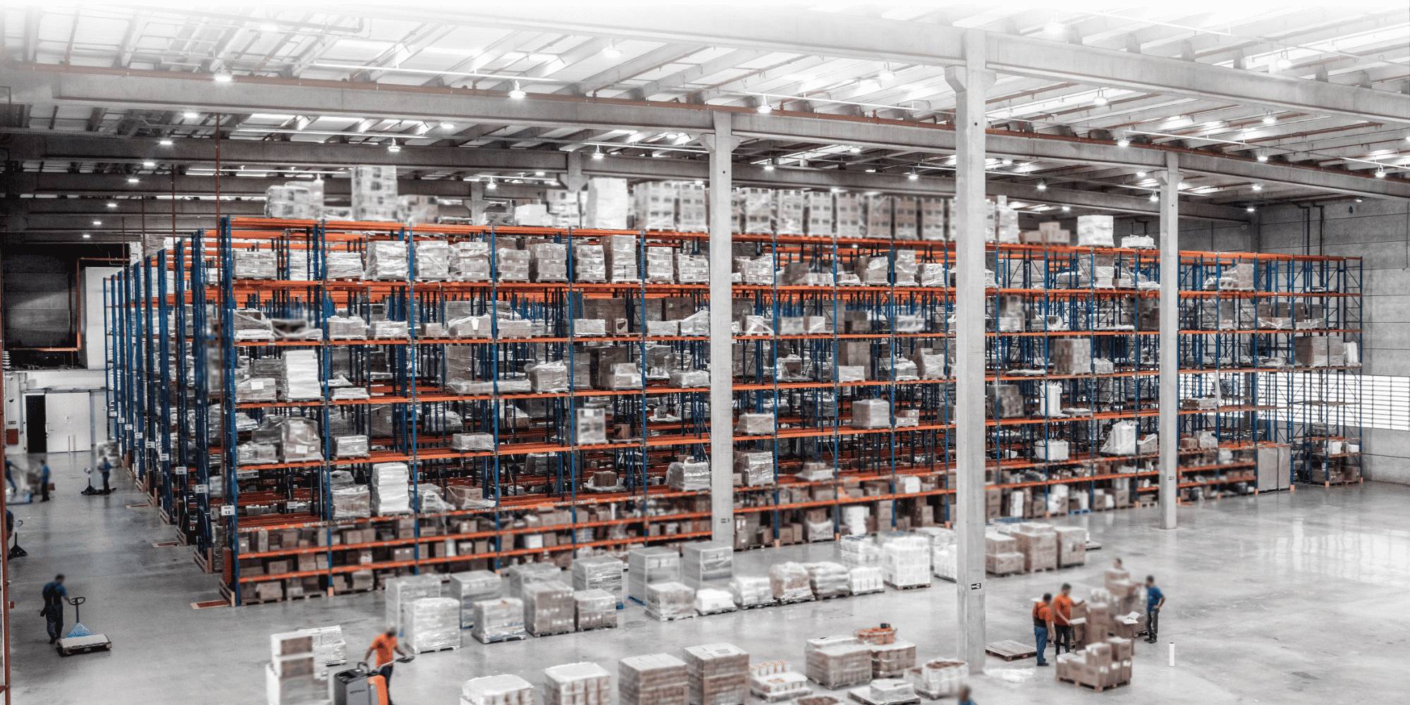 Una vista dall'alto di un ampio magazzino con alte scaffalature per pallet blu e arancioni su cui sono impilate merci imballate, operai che manovrano transpallet su un pavimento in cemento levigato e merce pallettizzata in attesa di spedizione sotto una brillante illuminazione industriale.