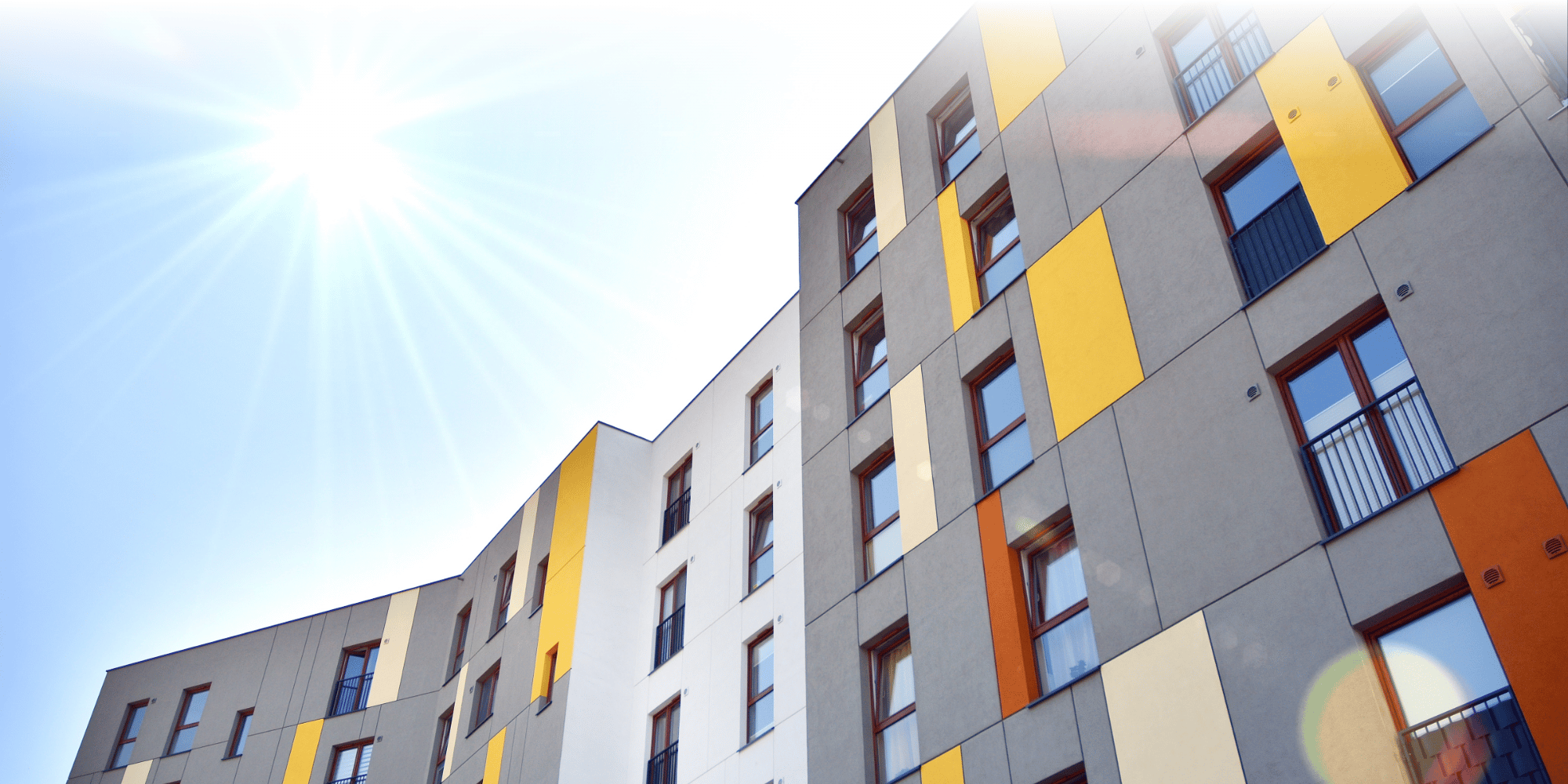 Low-angle view of a modern residential apartment block with a colourful grey, yellow, orange, and cream cladded facade, photographed against a bright sunny sky with lens flare.
