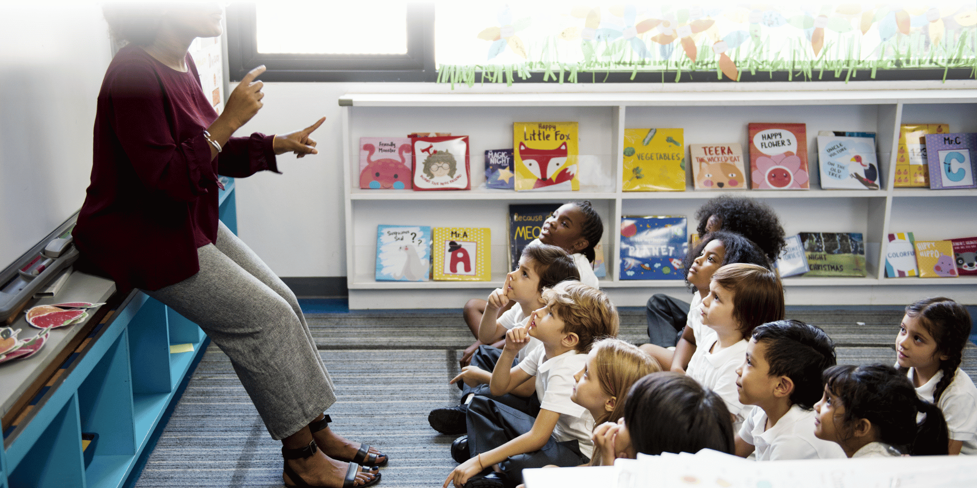 Un’insegnante sorridente che parla a dei bambini piccoli seduti sul pavimento in un’aula di scuola elementare decorata con colori vivaci.