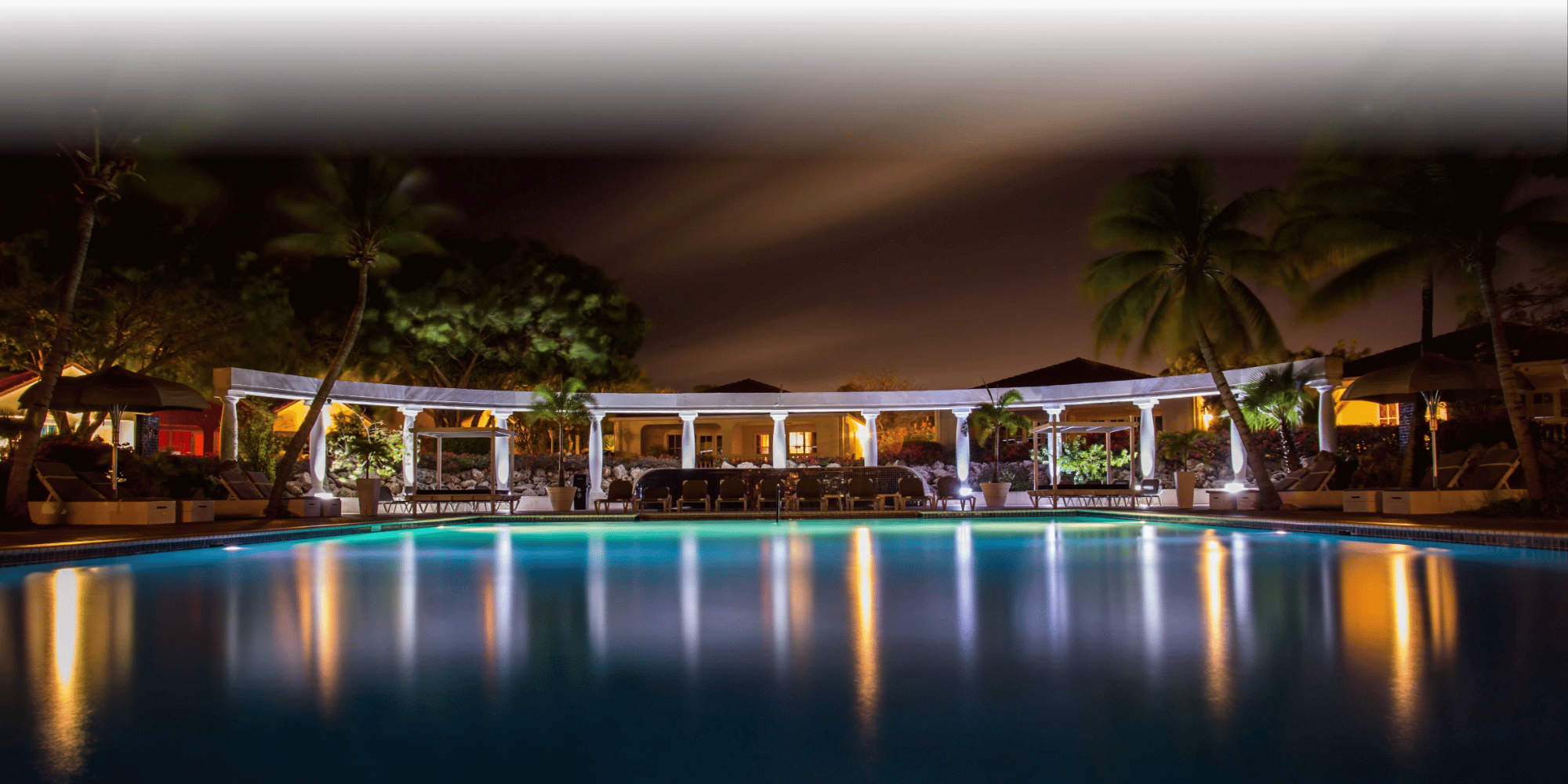 Nighttime view of a luxury resort pool illuminated with warm lights, framed by white colonnades, palm trees, and poolside loungers, with the lights reflecting on the calm turquoise water under a dark sky.