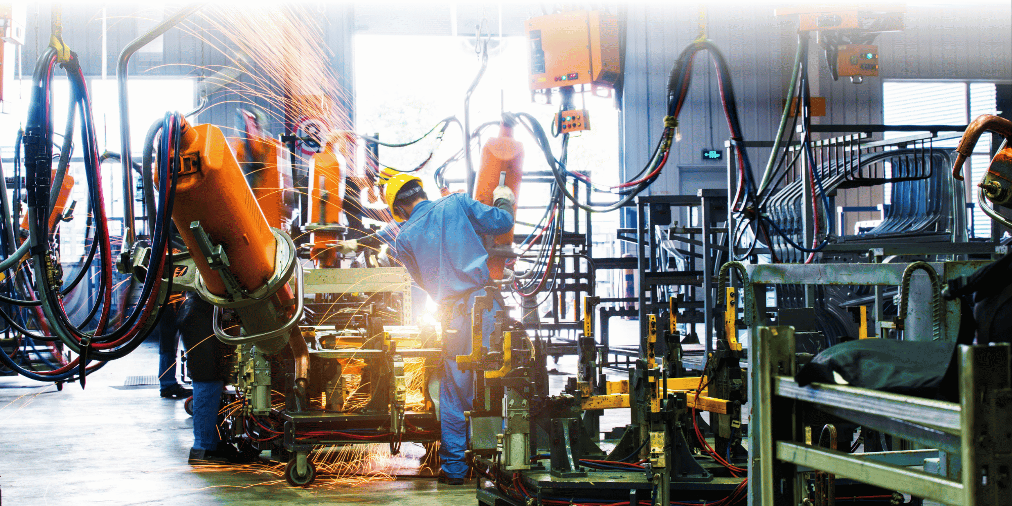 A worker in blue coveralls and a hard hat welds metal parts on a factory production line, surrounded by orange robotic arms, sparks, and manufacturing equipment.