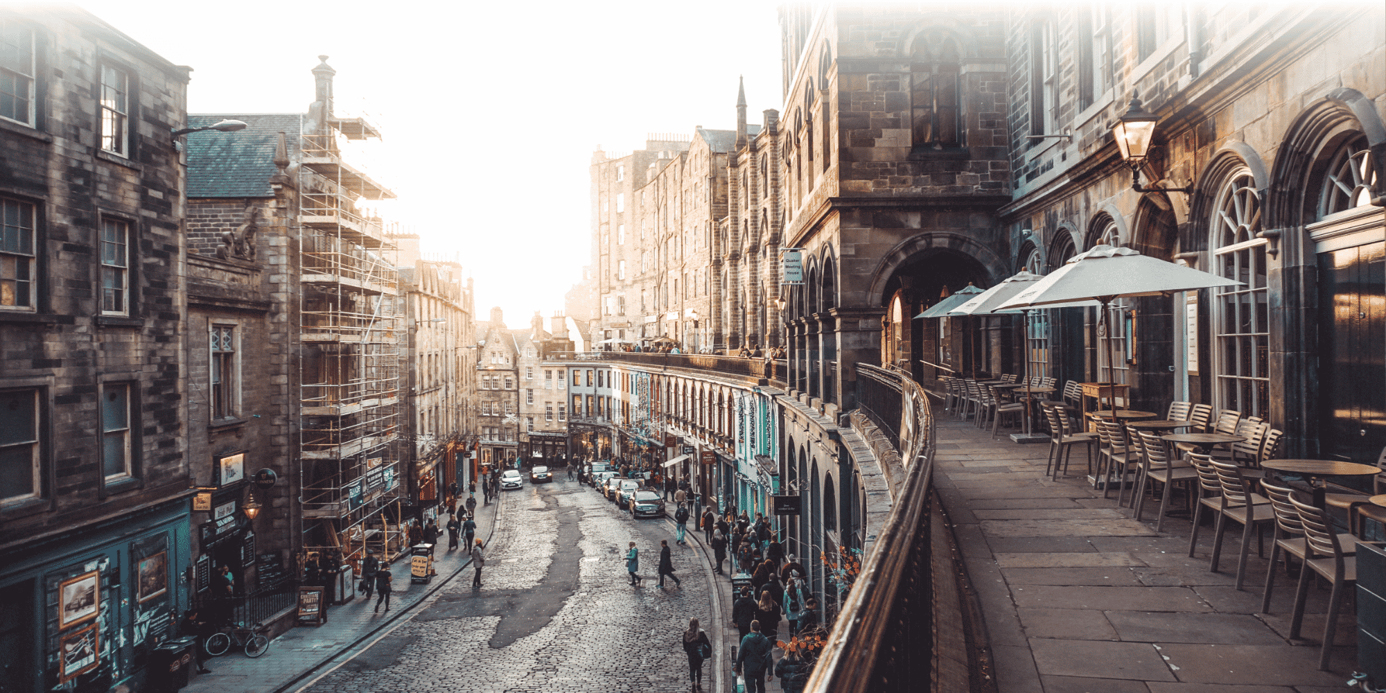 High-angle view of busy cobbled Edinburgh Old Town street, with colourful shops, pedestrians, and a café terrace with parasols beside a stone building.