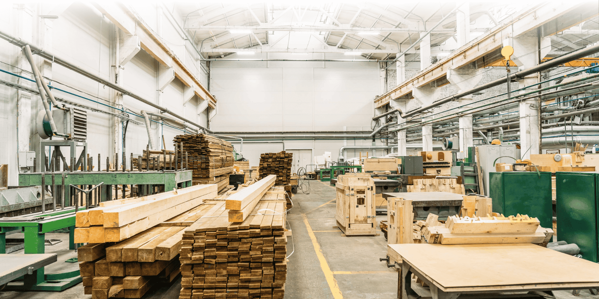 Capannone industriale con assi di legno impilate su un pavimento in cemento, affiancato da macchine per la lavorazione del legno sotto un tetto con struttura in acciaio dotato di binari per gru.
