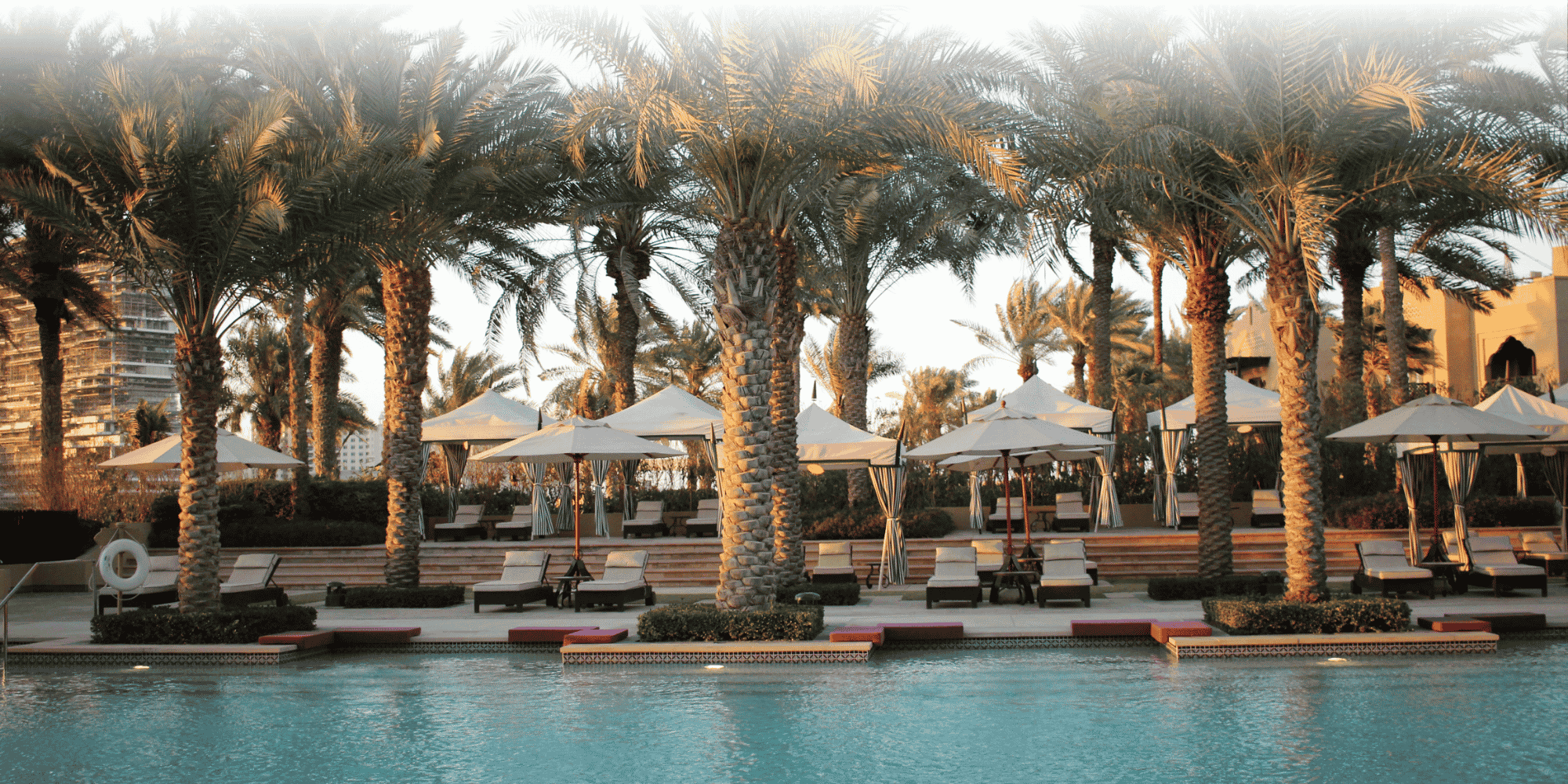 Turquoise pool bordered by date palms, with sun loungers, white parasols and pink poolside platforms in warm evening light.