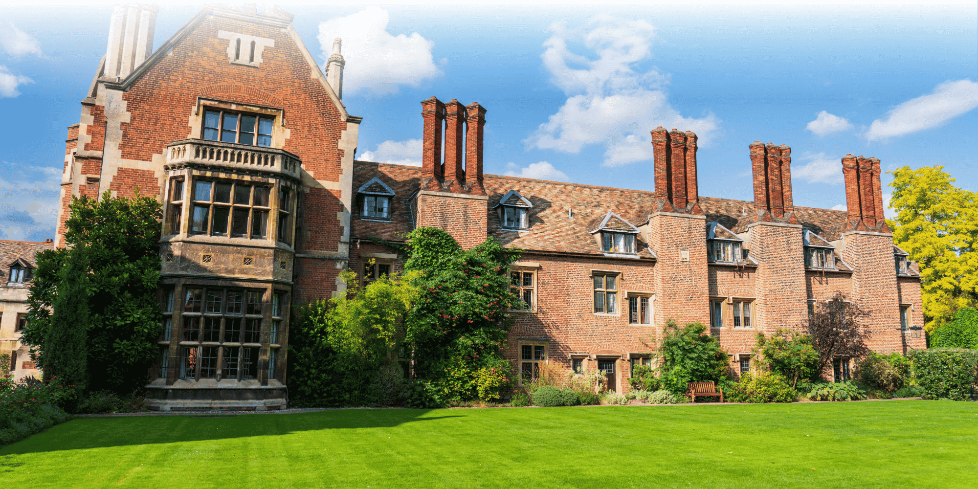 Grand Tudor red-brick building with ornate chimneys, mullioned windows and bay turret, set against manicured lawns and gardens under a blue sky.