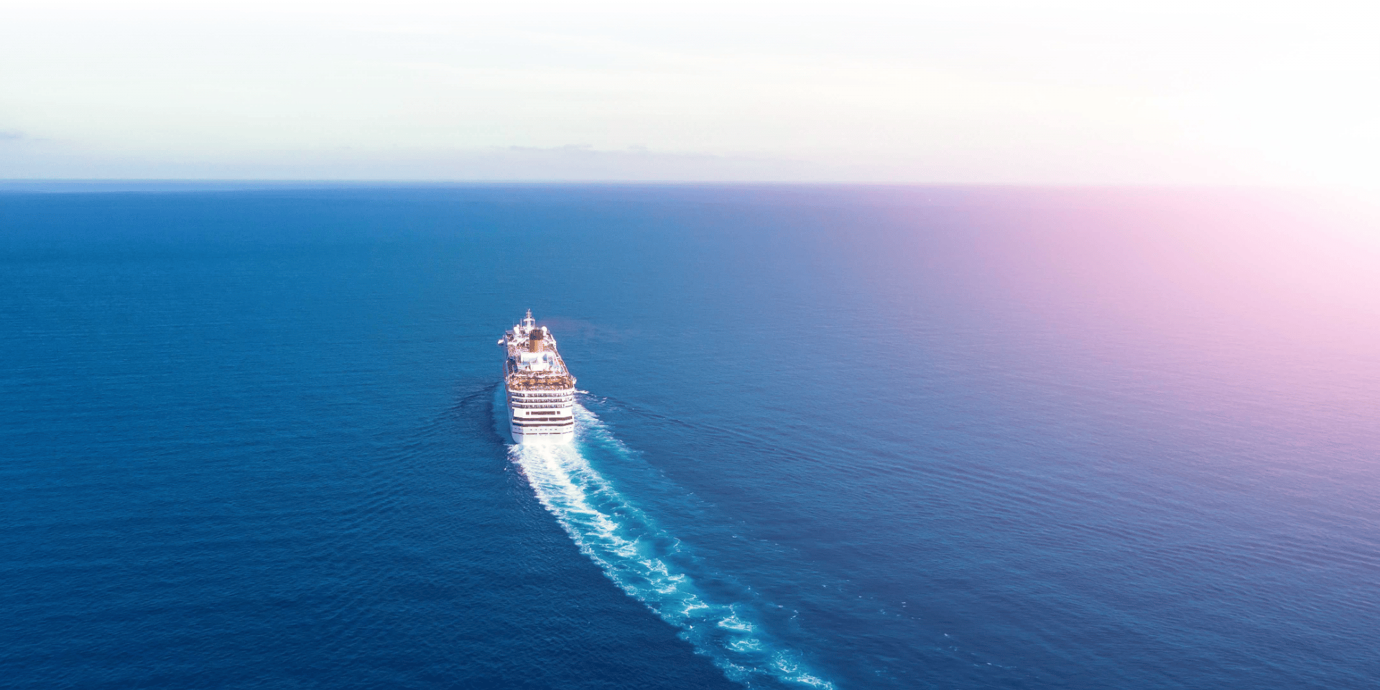Aerial view of a cruise ship sailing across open blue ocean, leaving a white wake trail beneath a hazy sky.Aerial view of a cruise ship sailing across open blue ocean, leaving a white wake trail beneath a hazy sky.