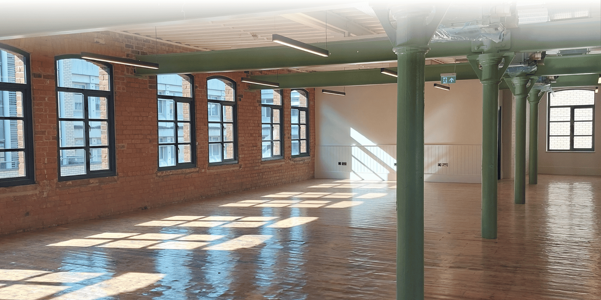 A bright, empty loft-style office space with exposed brick walls, tall arched windows, polished wood flooring, and green cast-iron columns and beams supporting the ceiling.