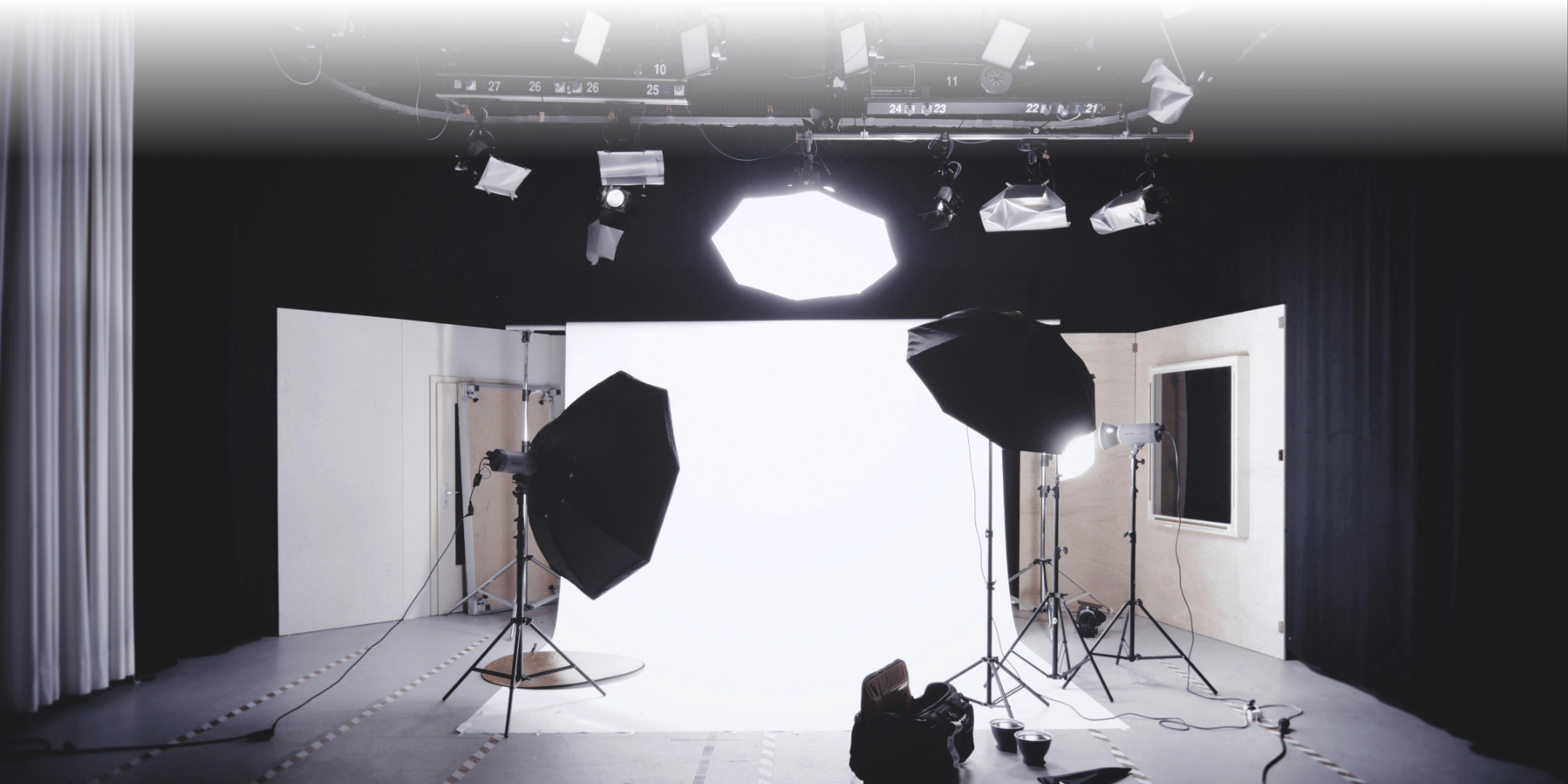 A professional photography studio with a white seamless backdrop, softbox lighting rigs on stands, an overhead ceiling-mounted lighting grid, and camera equipment on the floor.