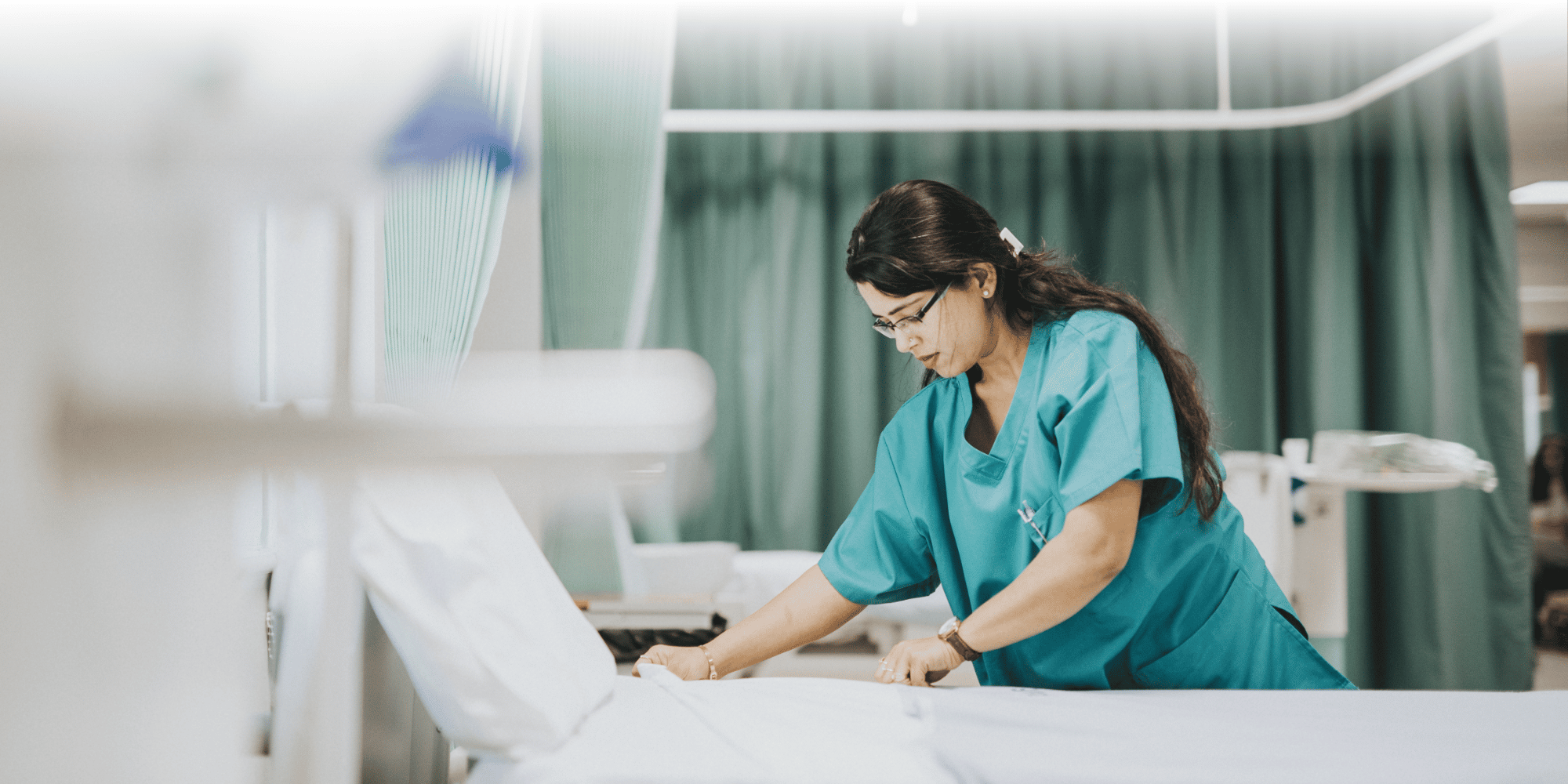 Un'infermiera in camice color verde acqua che rifà un letto d'ospedale con lenzuola bianche, con tende verde chiaro che garantiscono la privacy sullo sfondo.