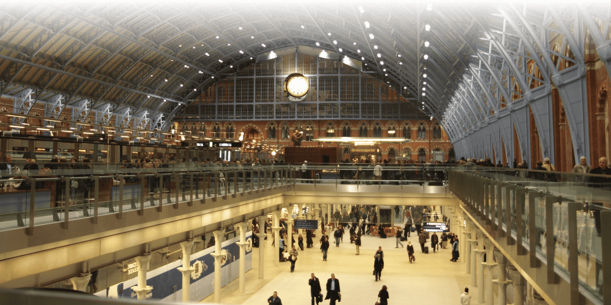 Interior of St Pancras railway station, showing its vaulted glass-and-iron roof, a large clock, upper-level shops and bars, and travelers on the concourse below.