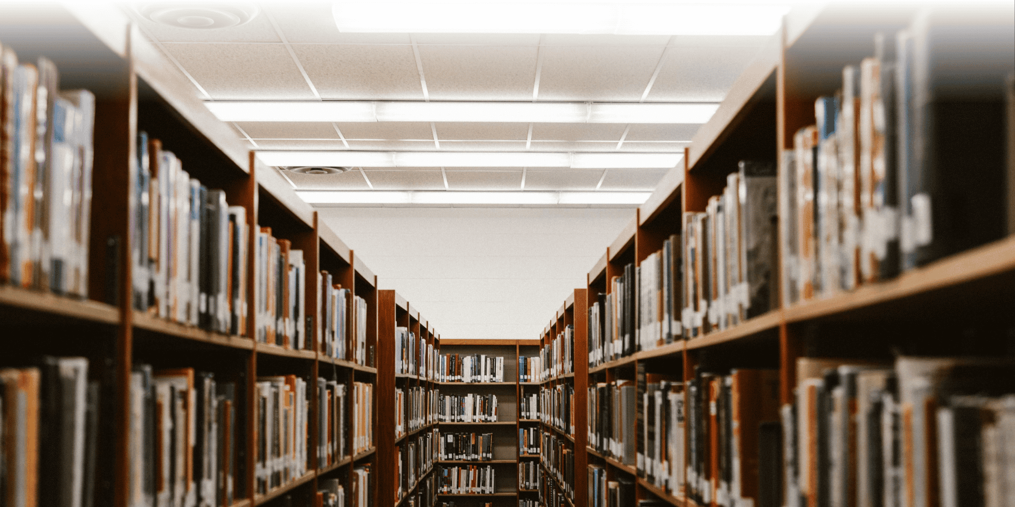 Library aisle lined with tall wooden bookshelves, with fluorescent strip lighting and a smoke detector on the ceiling.