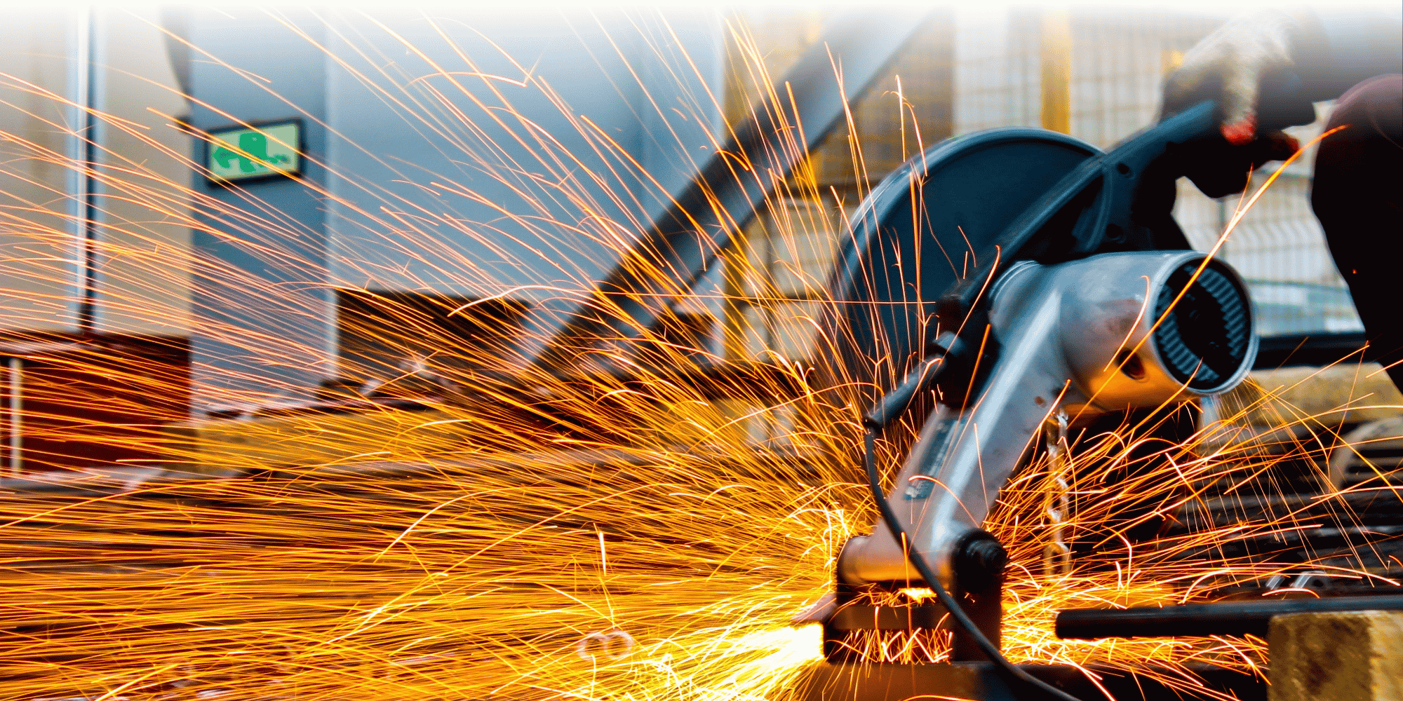 A gloved worker uses an angle grinder on metal at an industrial site, sending a dramatic shower of bright orange sparks across the foreground.