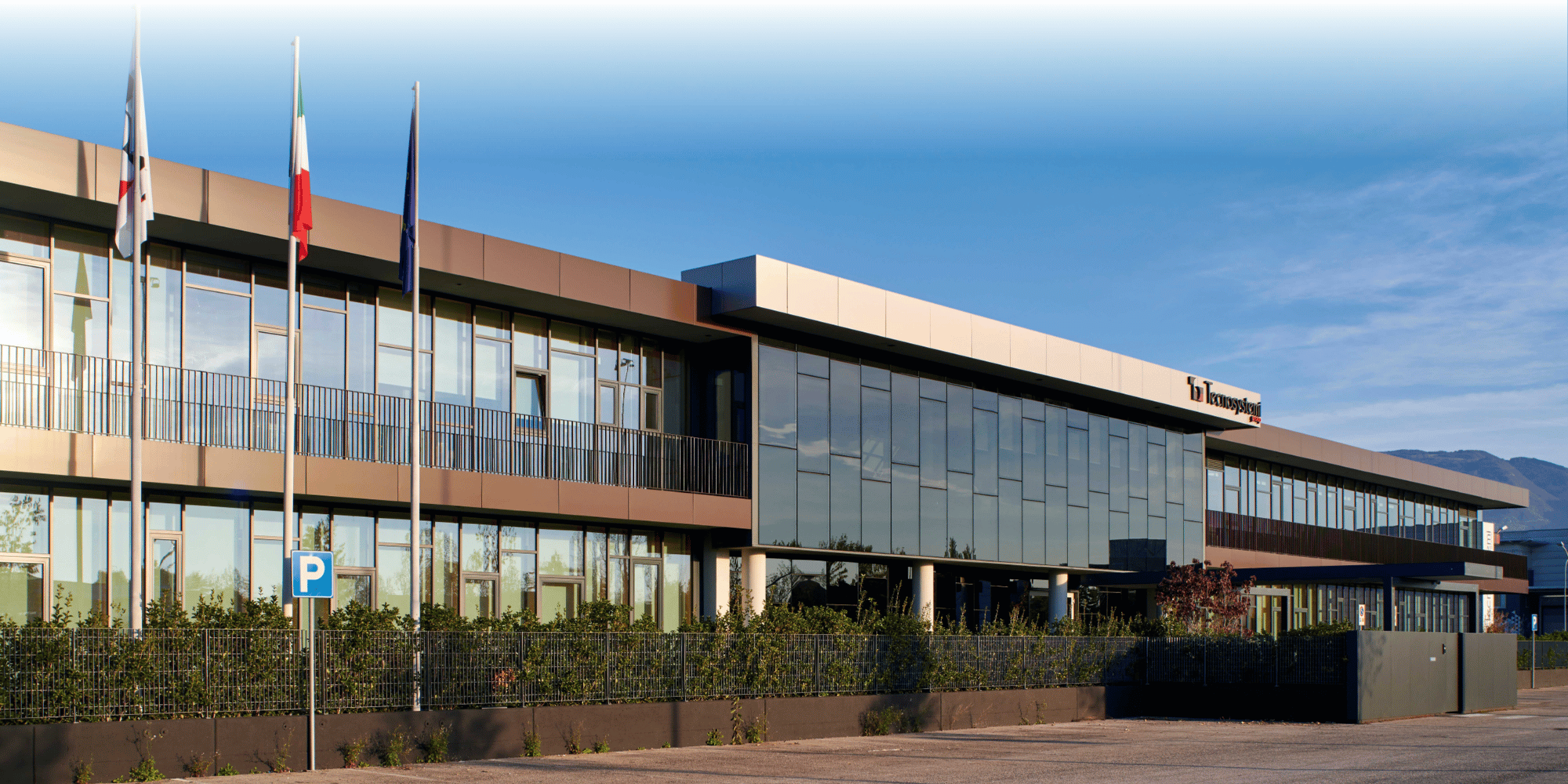 Modern two-story Tecnosystemi office building with glass facade, balconies, and flagpoles, set against a clear blue sky with distant mountains visible.