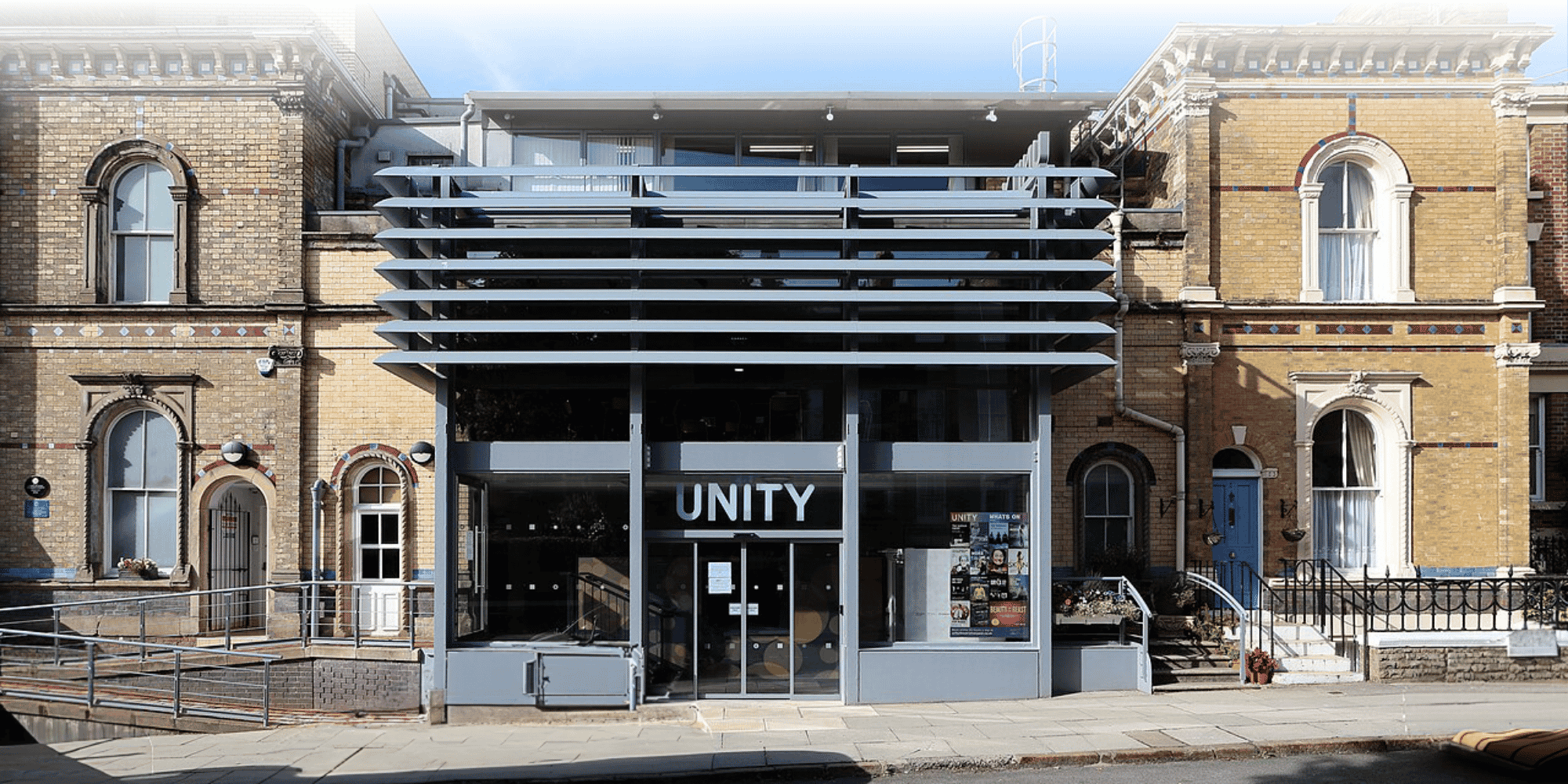 Street-level view of Unity building with contemporary grey glass façade and horizontal louvres, flanked by Victorian yellow brick buildings.