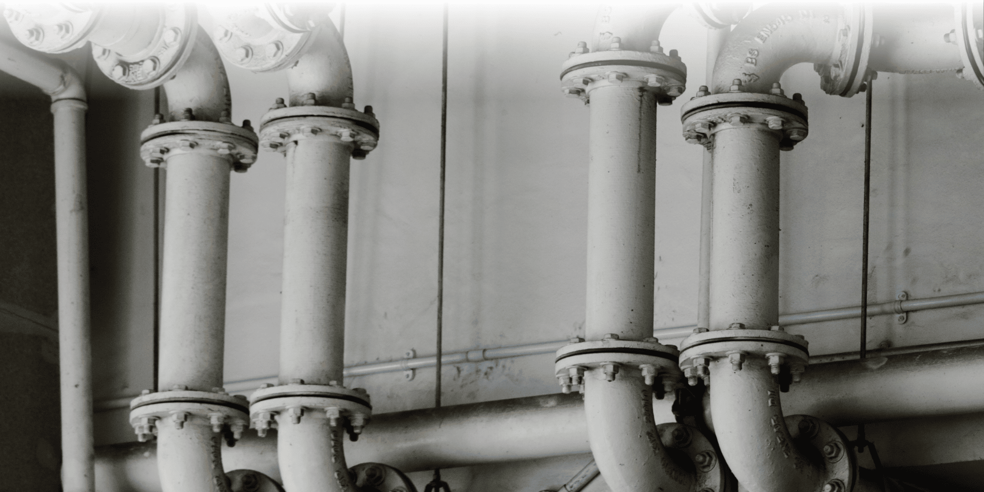 Black-and-white close-up of industrial pipework, showing several vertical pipes connected with bolted flange joints against a wall.