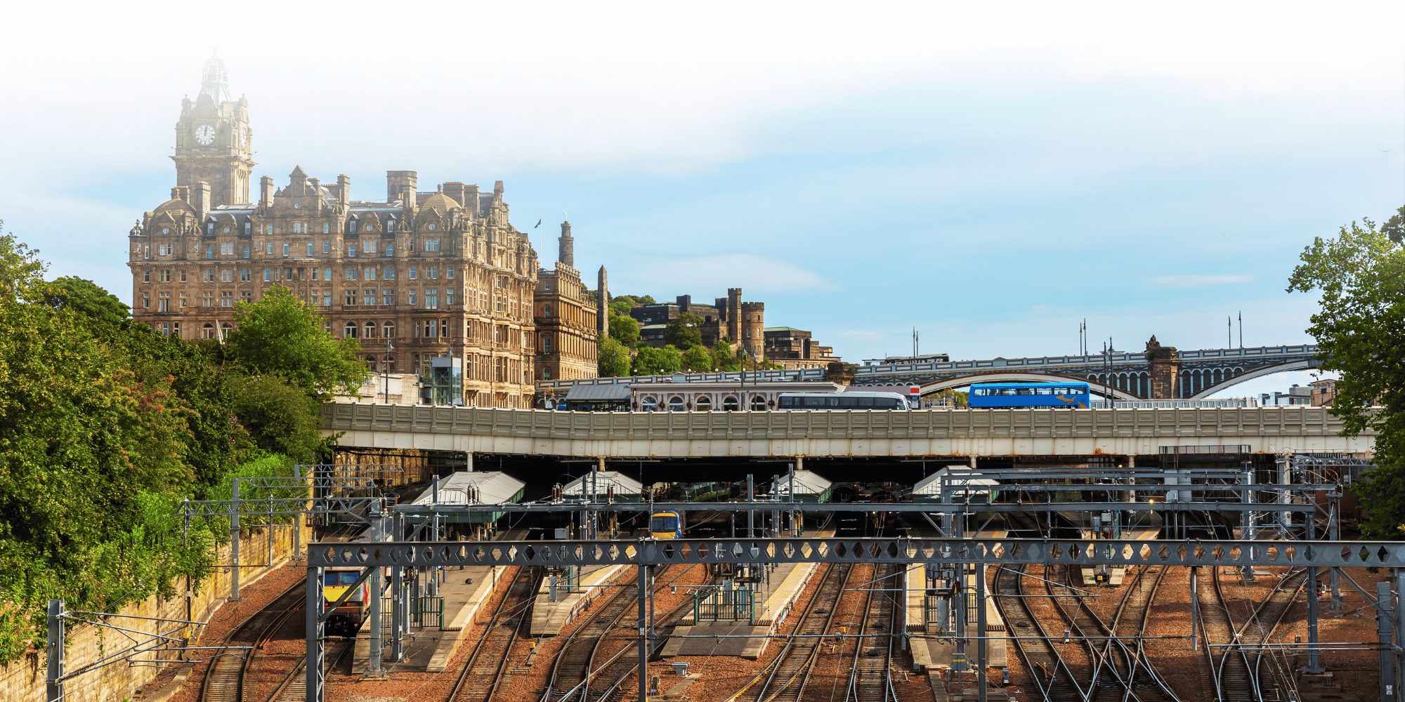 Aerial view of Edinburgh Waverley station's platforms and rail tracks, with the historic Balmoral Hotel clock tower and Edinburgh Castle visible in the background.