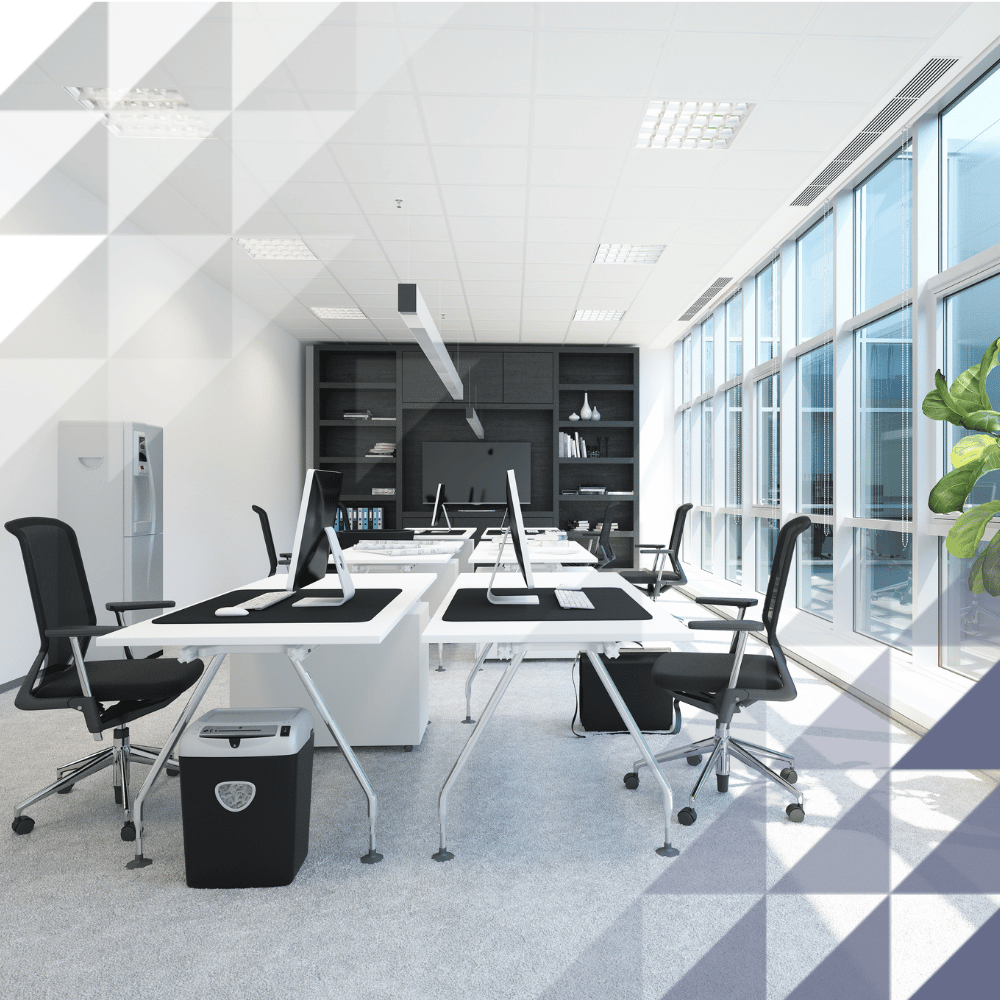 Modern open-plan office space with rows of desks, computer monitors, ergonomic chairs and large floor-to-ceiling windows providing natural light. The bright workspace features contemporary furnishings, shelving units and a clean, professional environment.