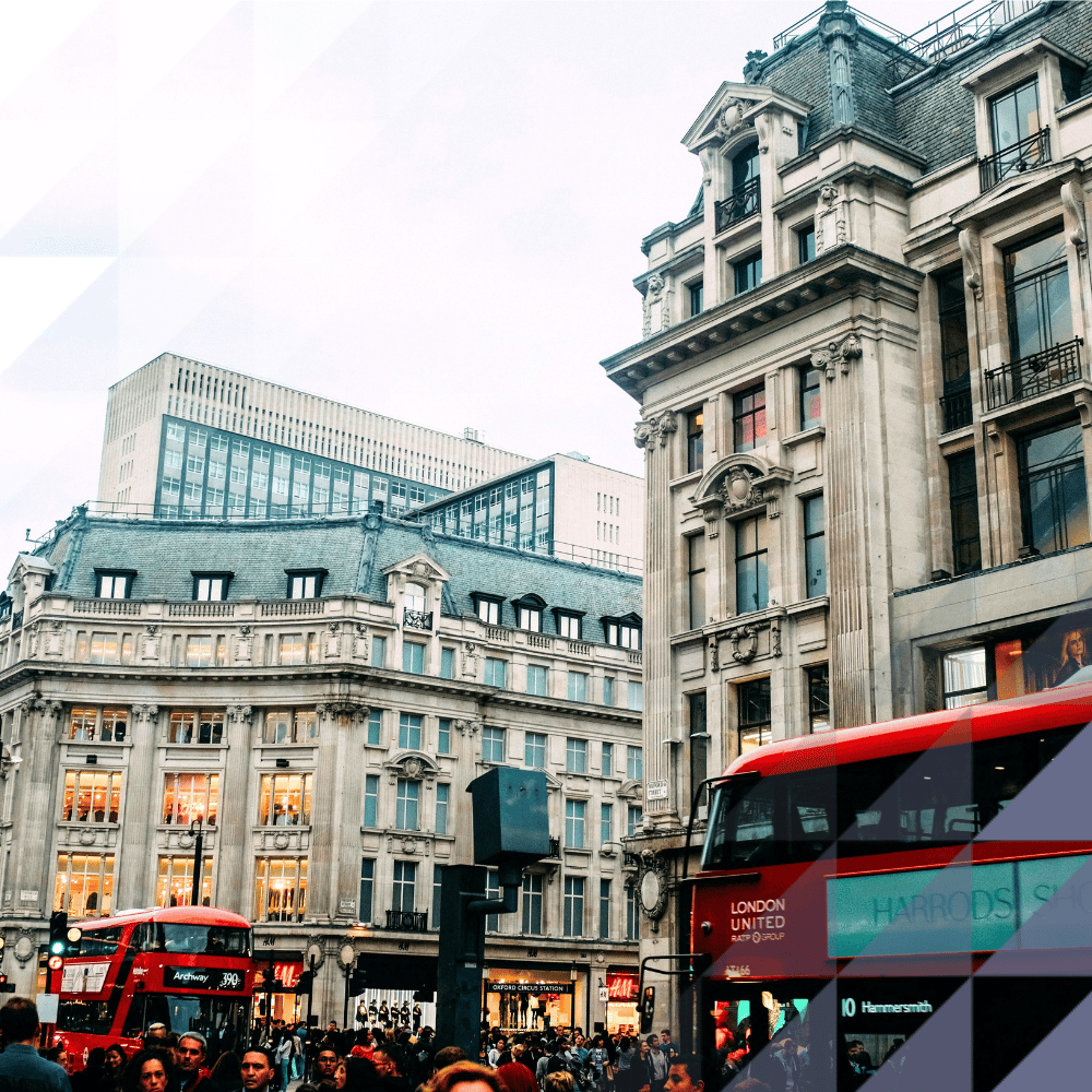Busy street scene at Oxford Circus, London, featuring historic and modern commercial buildings, crowds of pedestrians, and two iconic red double-decker buses travelling along Oxford Street.