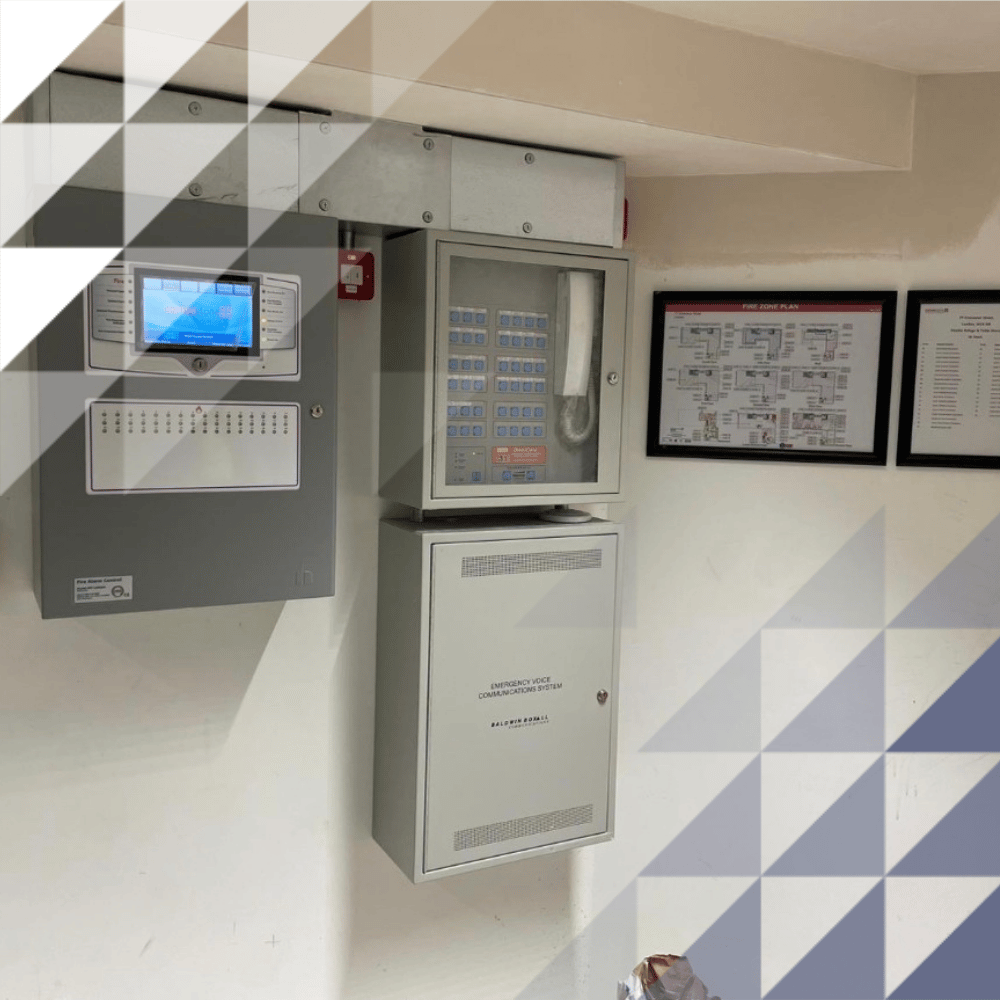 Wall-mounted fire alarm control panels with a touchscreen display, a manual call point, a communications cabinet, and framed fire escape plans on a white wall.