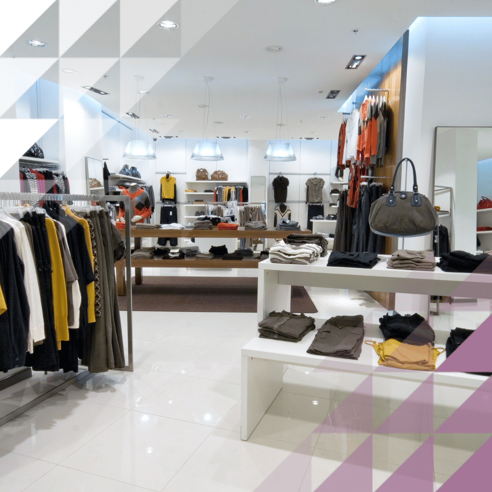Boutique di abbigliamento contemporaneo con capi appesi alle grucce, vestiti piegati e borse esposti sui tavoli, e scaffali illuminati sullo sfondo.
