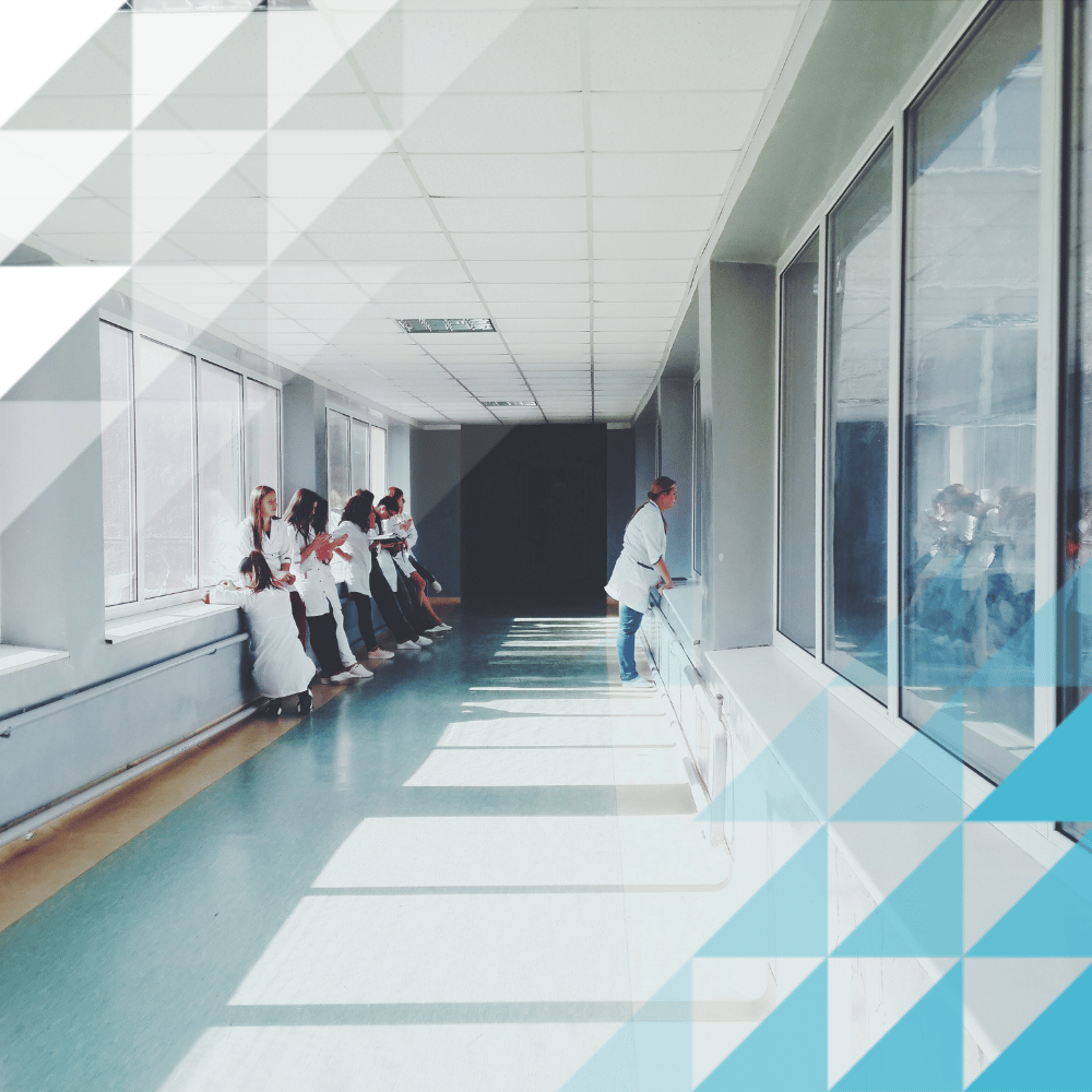Sunlit hospital hallway with a group of medical staff in white coats gathered along one wall and a lone clinician by a handrail opposite.