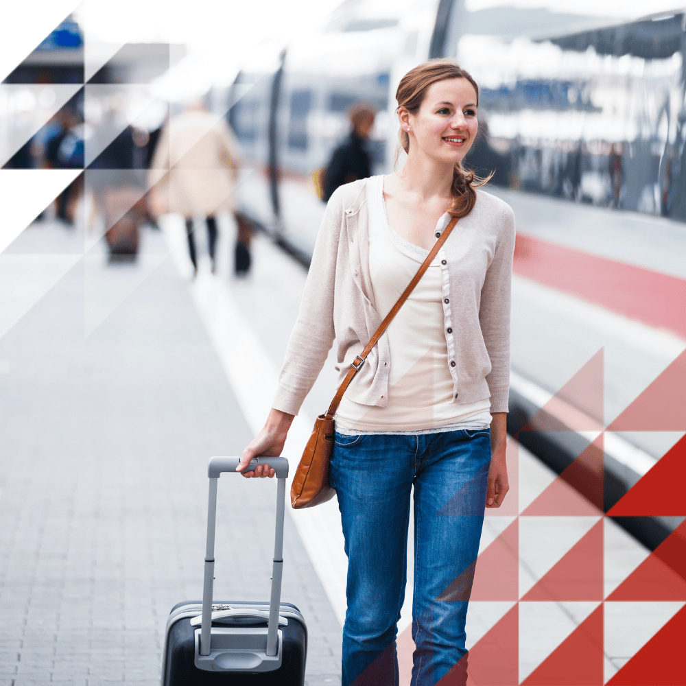 A traveller walks along a station platform with a suitcase beside a high-speed train, with other passengers visible in the background.