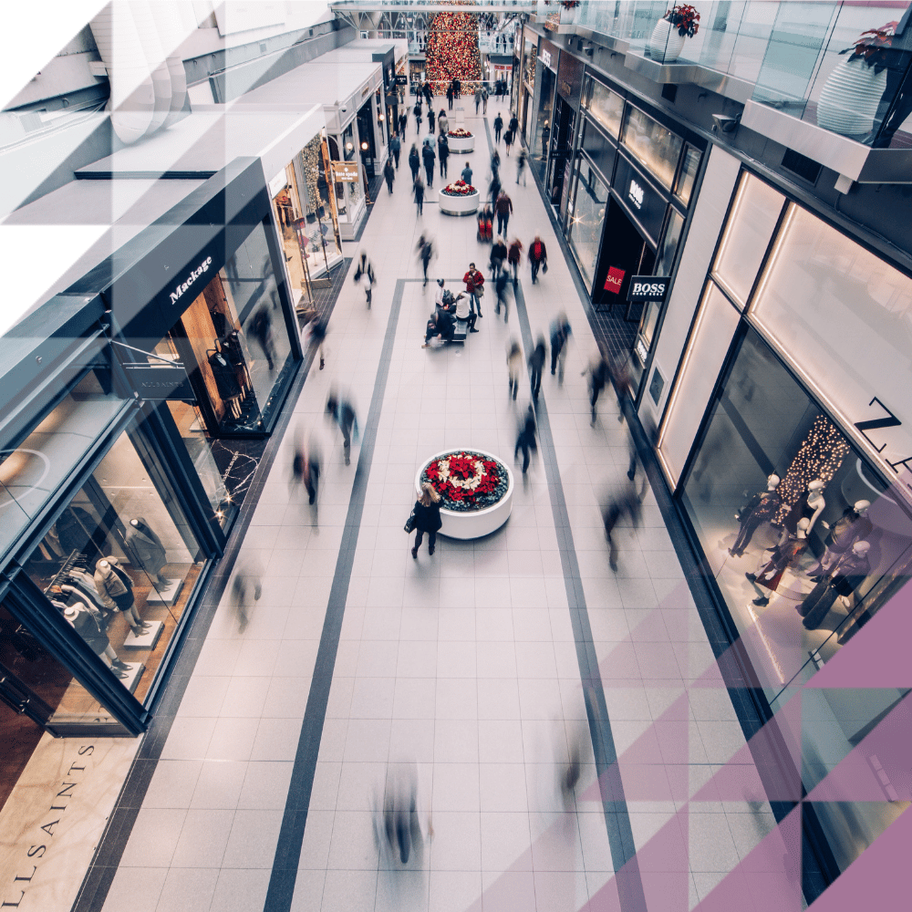 Aerial view of a busy shopping centre mall with shoppers moving through a wide concourse lined with retail stores, with a festive floral display at the centre.
