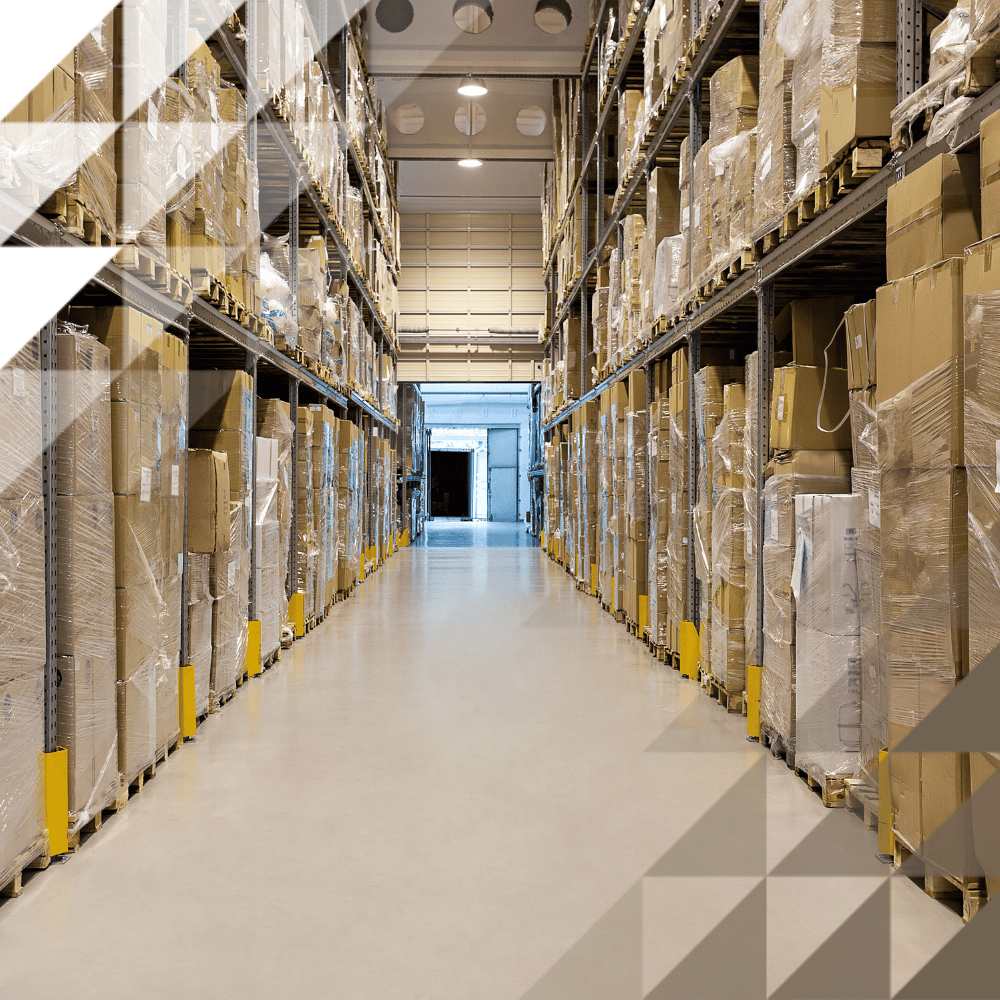 A narrow warehouse aisle flanked by shelving stacked with cardboard boxes and wrapped pallets, leading to a loading bay door at the far end.