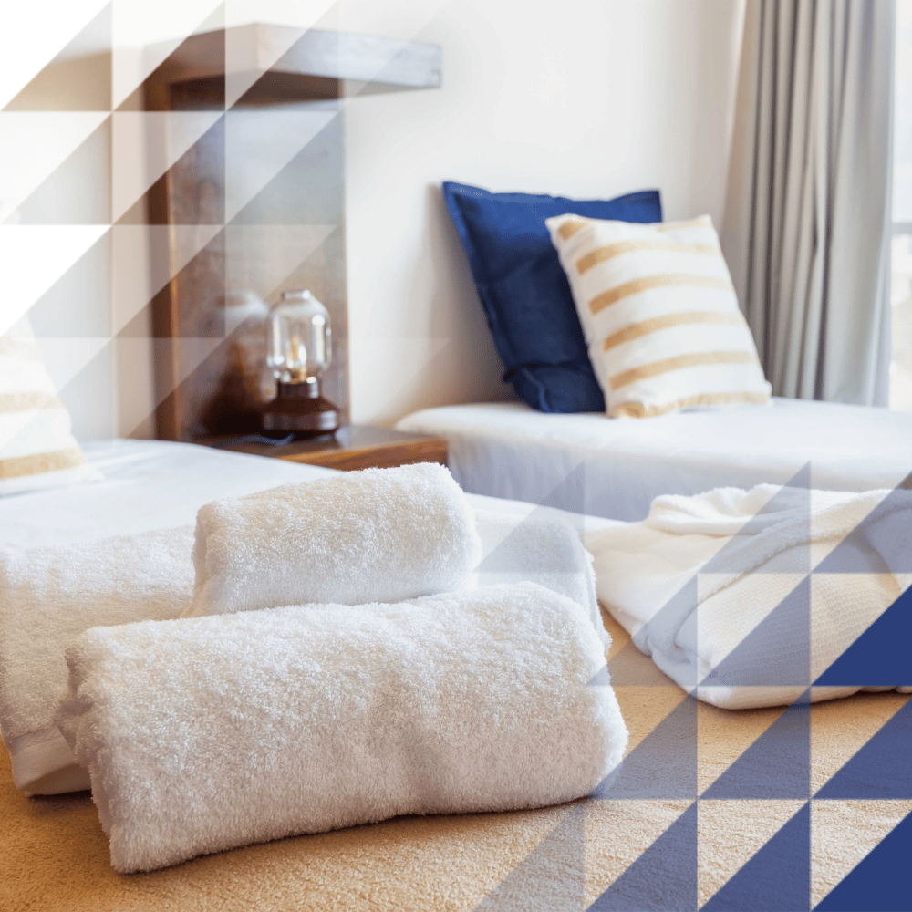 wo beds in a hotel room with towels and sheets layed on top. Both beds contain a beige cover, stripey and dark blue pillows