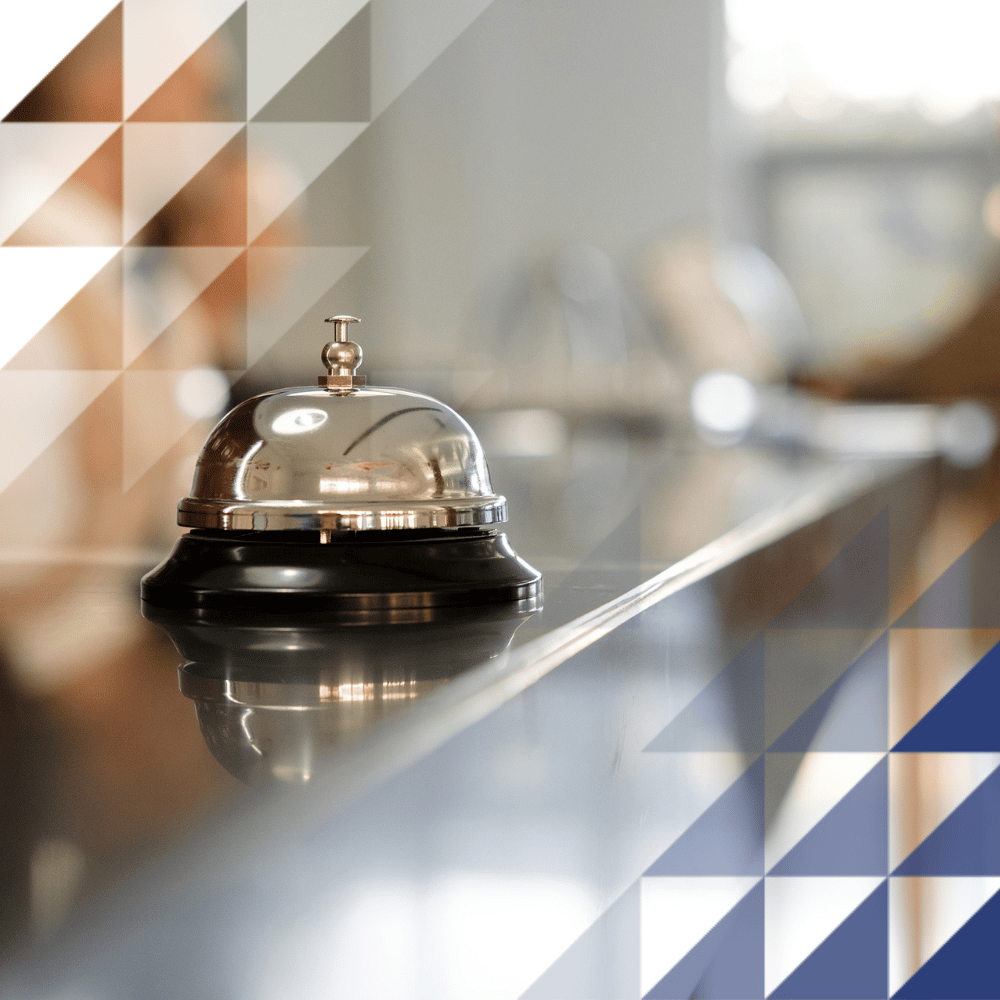 A polished service bell on a hotel reception desk, with blurred staff and guests in the background.