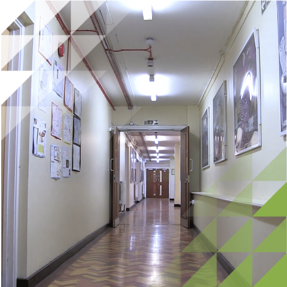 School corridor with cream walls displaying children's artwork, a red fire alarm pipe along the ceiling, and fluorescent strip lighting.