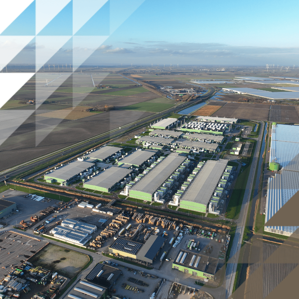 Bird's-eye view of industrial estate with rows of warehouse units, bordered by open fields, wind turbines and a solar farm under a clear sky.