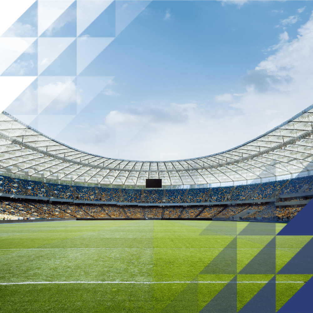 Ground-level view of a large football stadium with a green pitch, yellow and blue tiered seating, a curved canopy roof and a blue sky above.