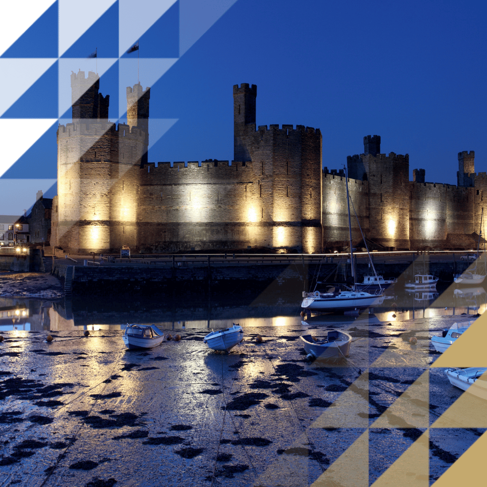 Caernarfon Castle's medieval sandstone walls and towers reflected in the harbour, with moored sailing boats under a dark night sky
