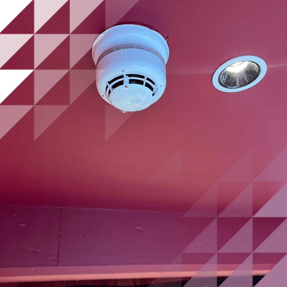 Un rilevatore di fumo bianco a soffitto accanto a un faretto da incasso, installato su un soffitto dipinto di rosa acceso all’interno di un locale commerciale o ricettivo.